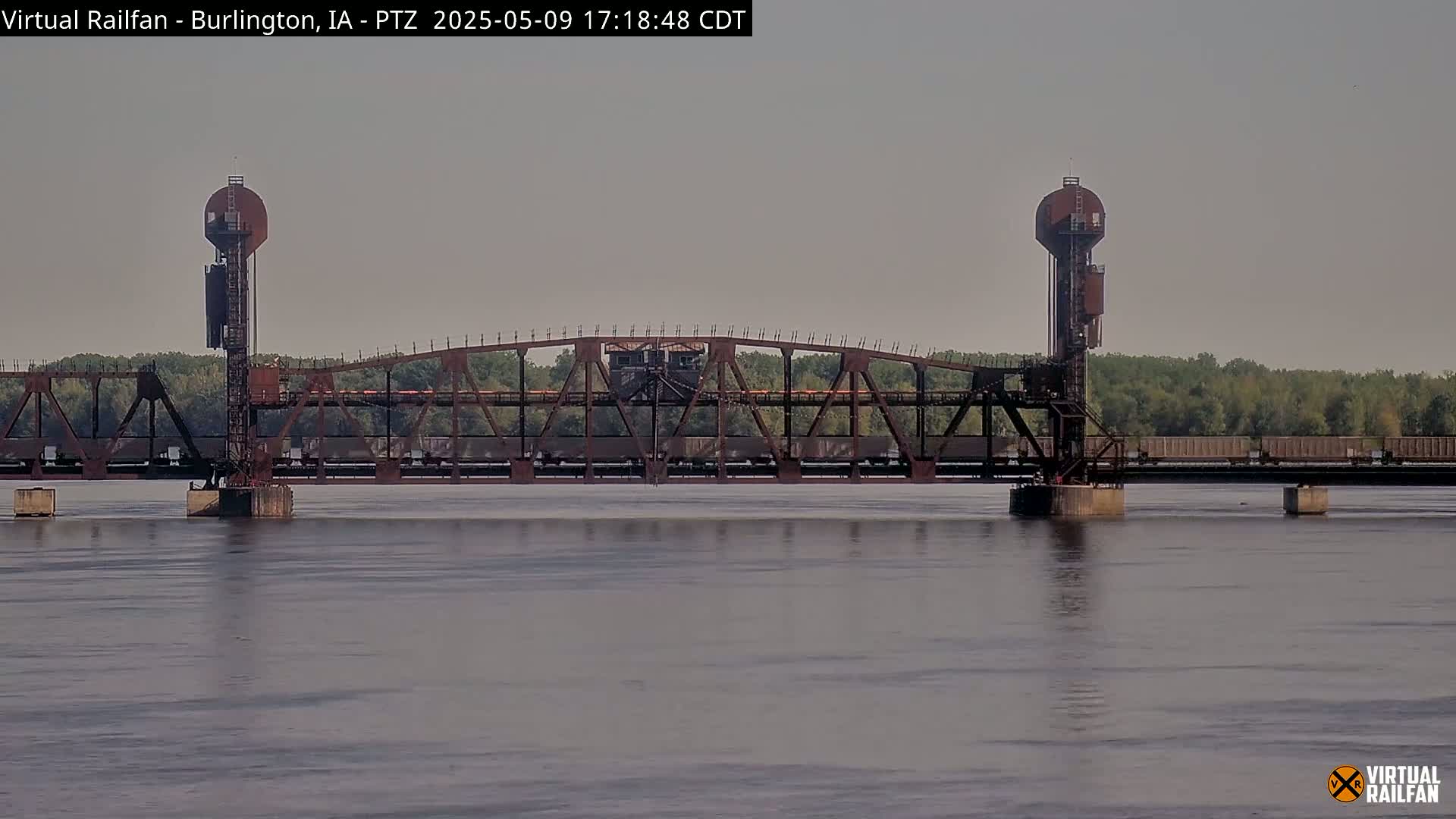 A rusty railroad drawbridge with a train on it spans a calm river under a hazy sky.