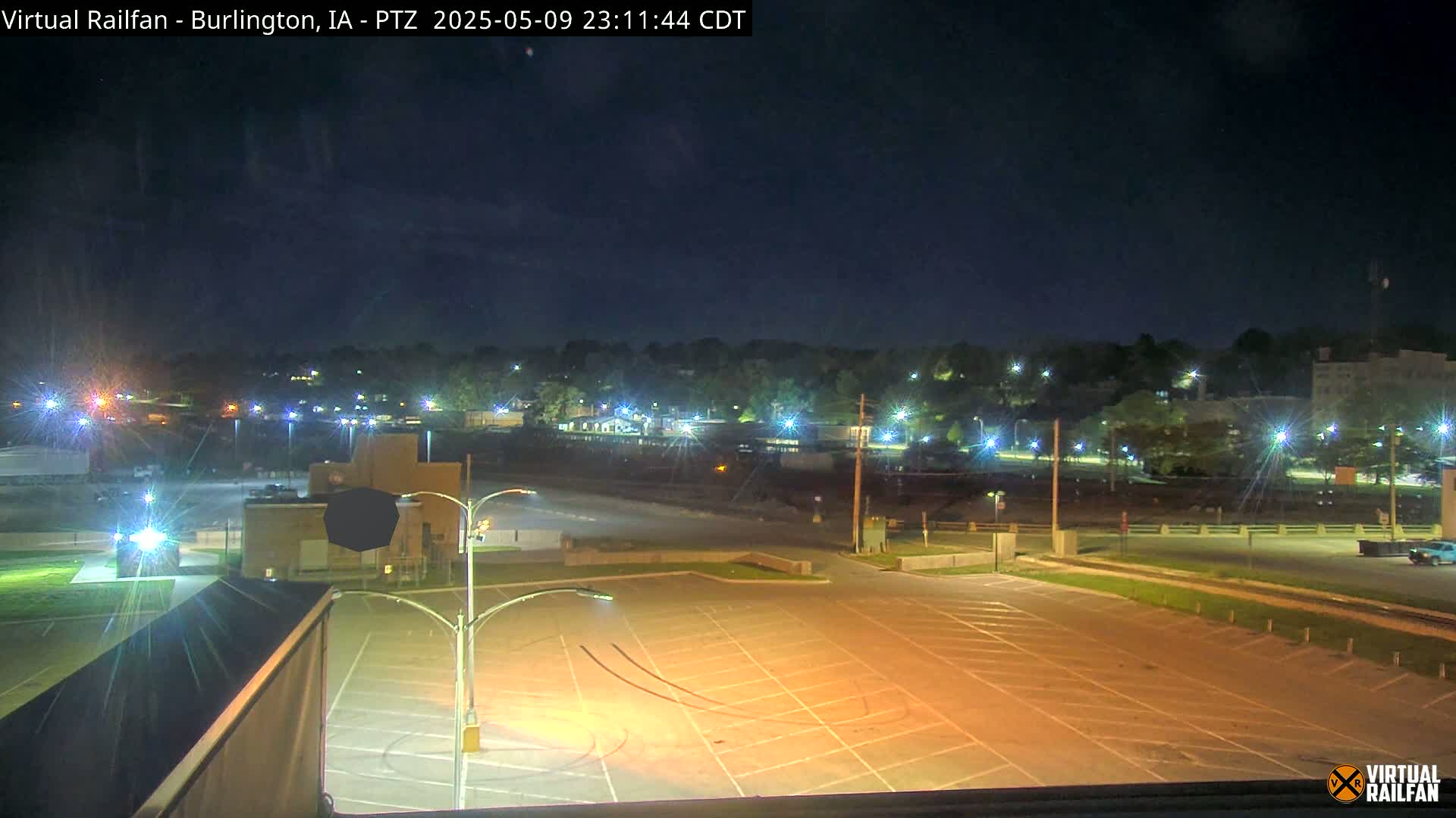 An empty, illuminated parking lot at night overlooks a tree-lined cityscape under a clear, dark sky.