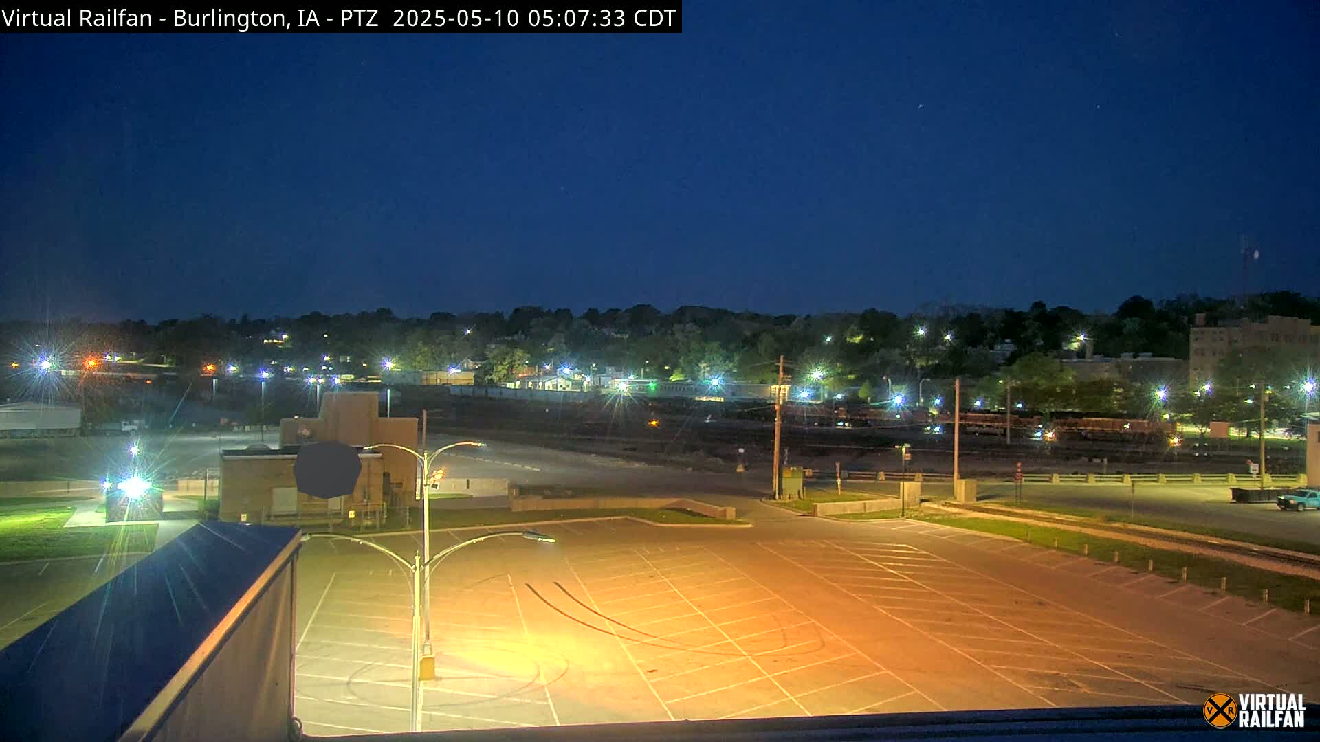 An empty, illuminated parking lot at night overlooks a railway yard and a treeline of buildings under a dark blue, clear sky.