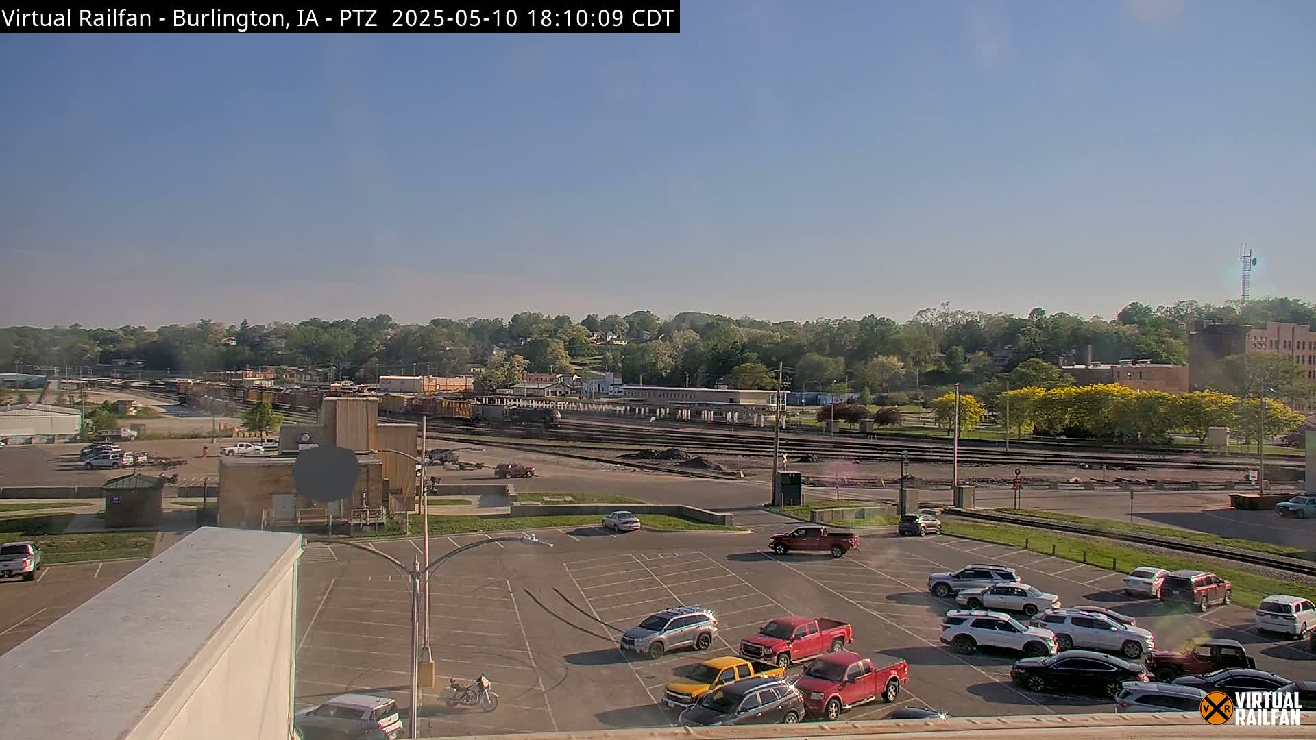 A long freight train sits in a rail yard under a clear blue sky, with several cars parked in a lot in the foreground.