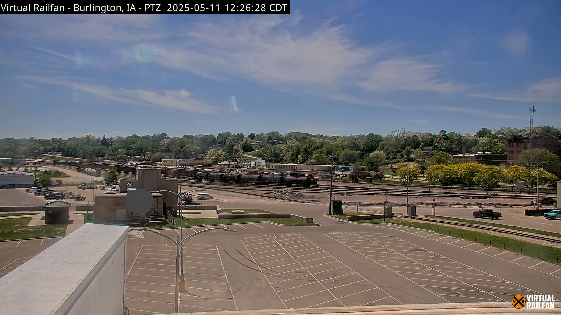 A mostly empty parking lot overlooks a railway yard filled with train cars under a partly cloudy, sunny sky.