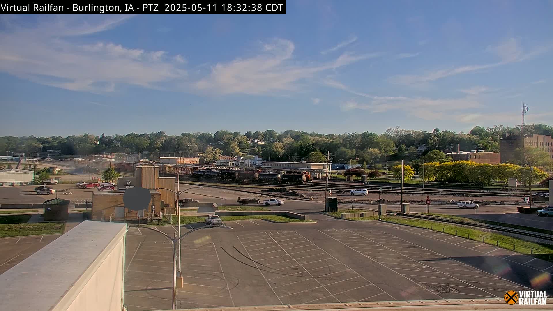 A mostly sunny day shows a large train yard with several rail cars and a few vehicles in a parking lot, surrounded by trees and buildings.