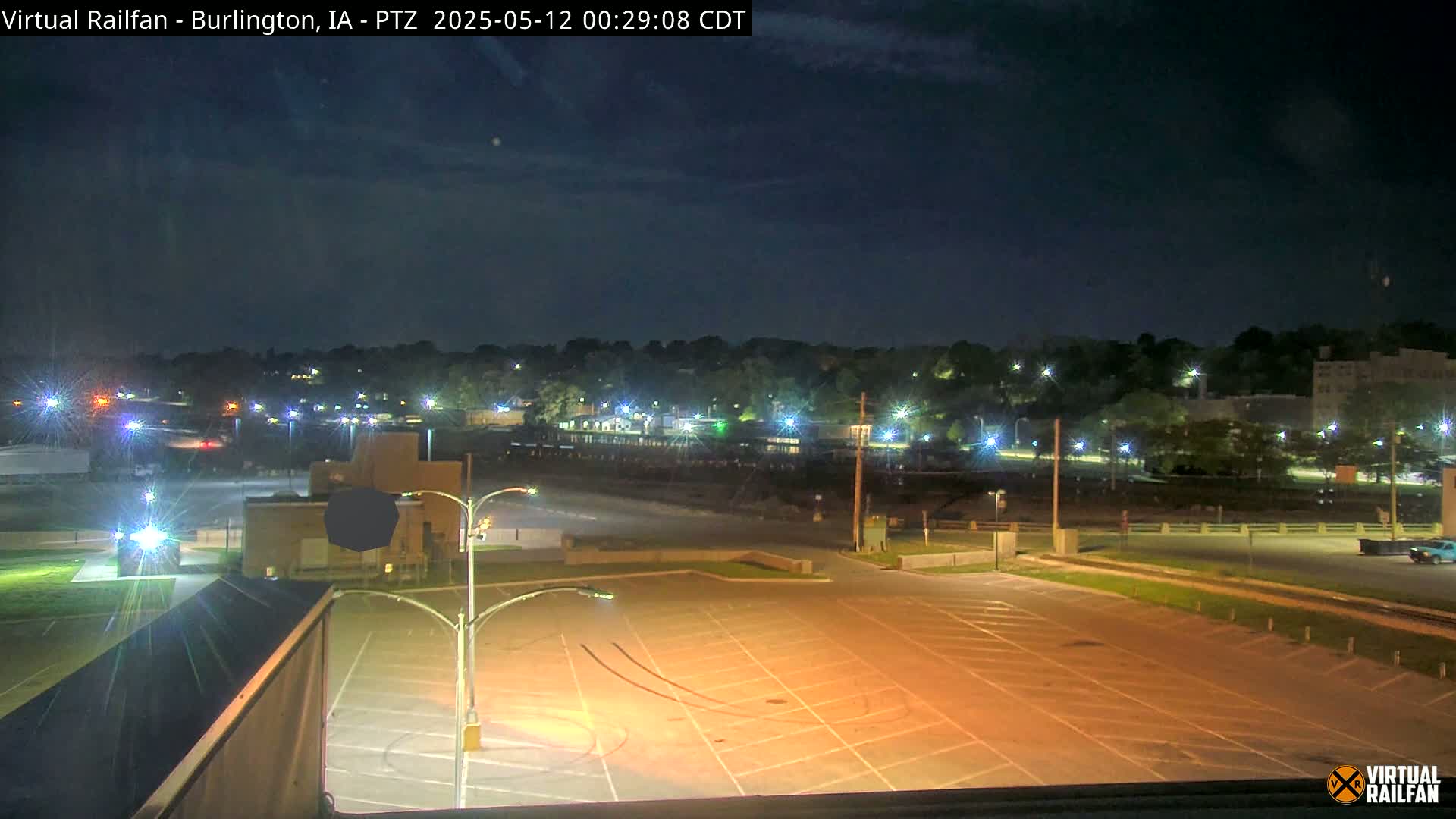 An empty, illuminated parking lot at night overlooks a tree-lined area with distant buildings and streetlights under a mostly clear sky.