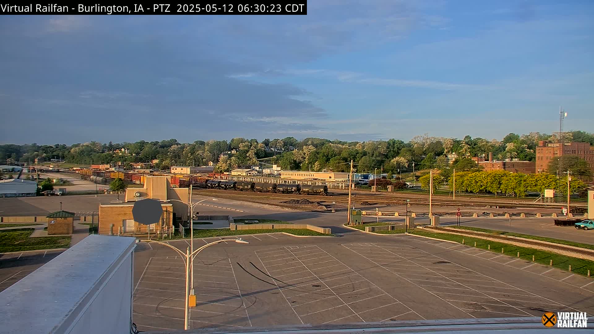 A mostly empty parking lot overlooks a rail yard with several train cars, and a tree-lined hill in the background under a partly cloudy, sunny sky.