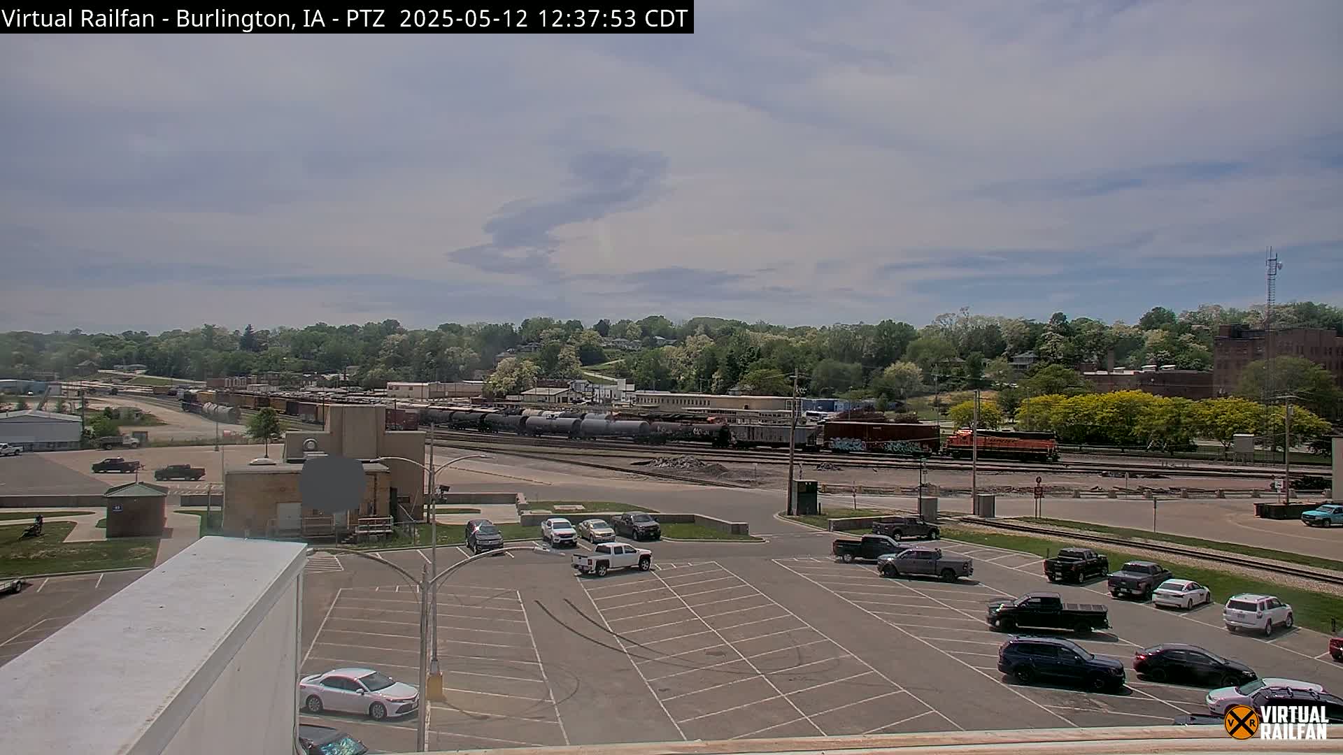 A partly cloudy day reveals a train yard with numerous railcars and a parking lot with several cars, all situated in front of a tree-lined hill.