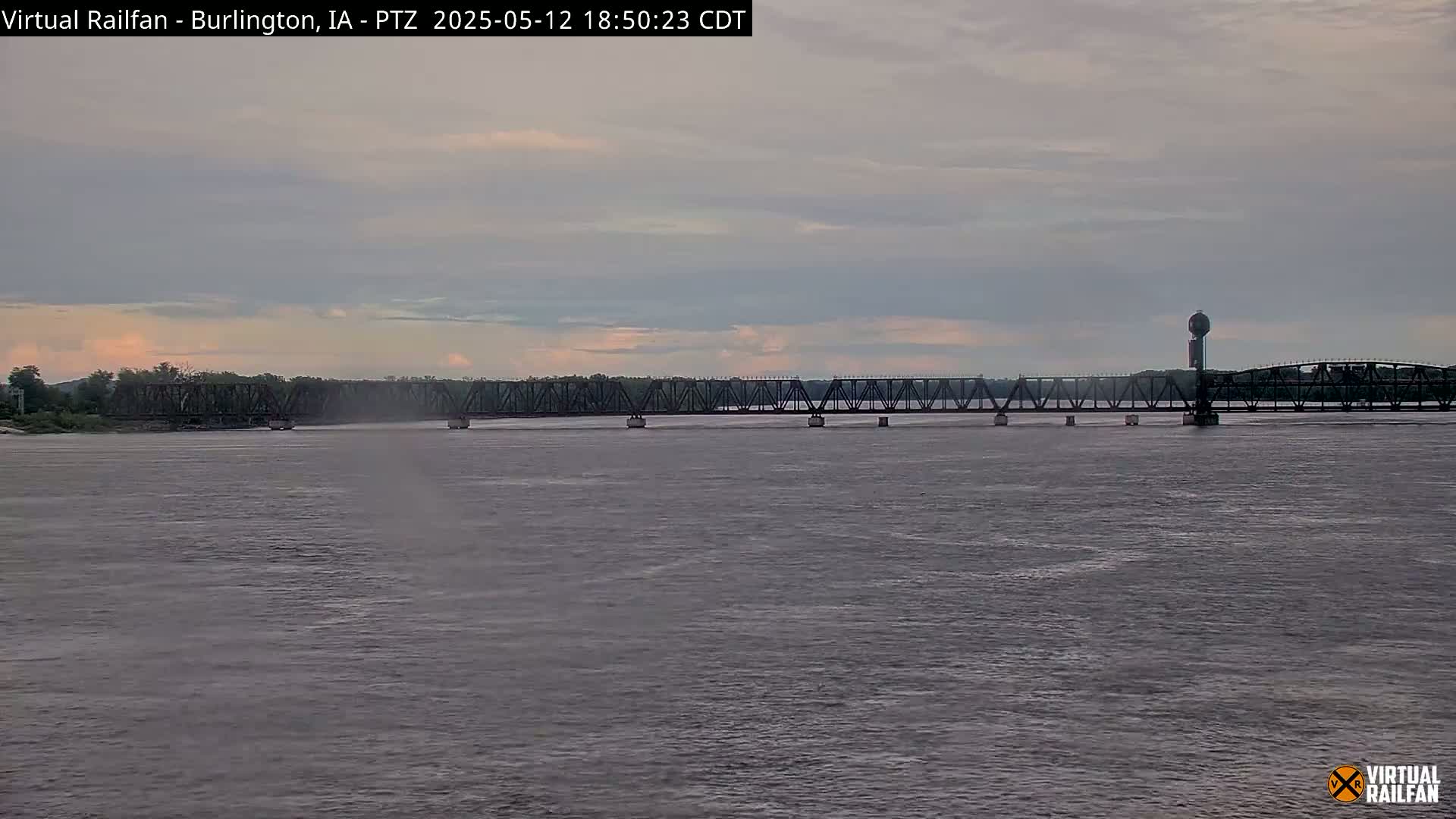 A long, metal truss bridge spans a wide river under an overcast sky.