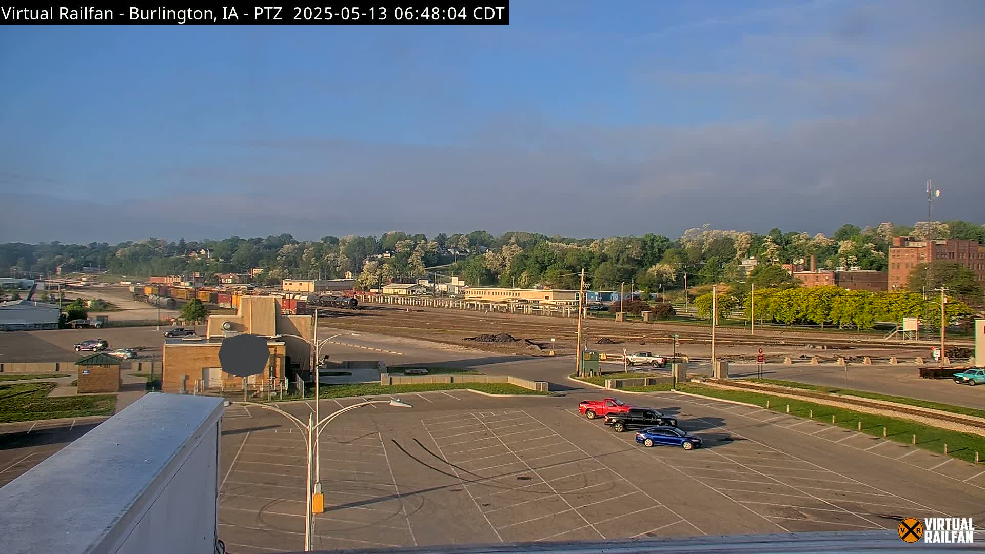 A mostly clear, bright sky overlooks a train yard with several rail cars, a parking lot with a few vehicles, and a tree-lined hill in the background.