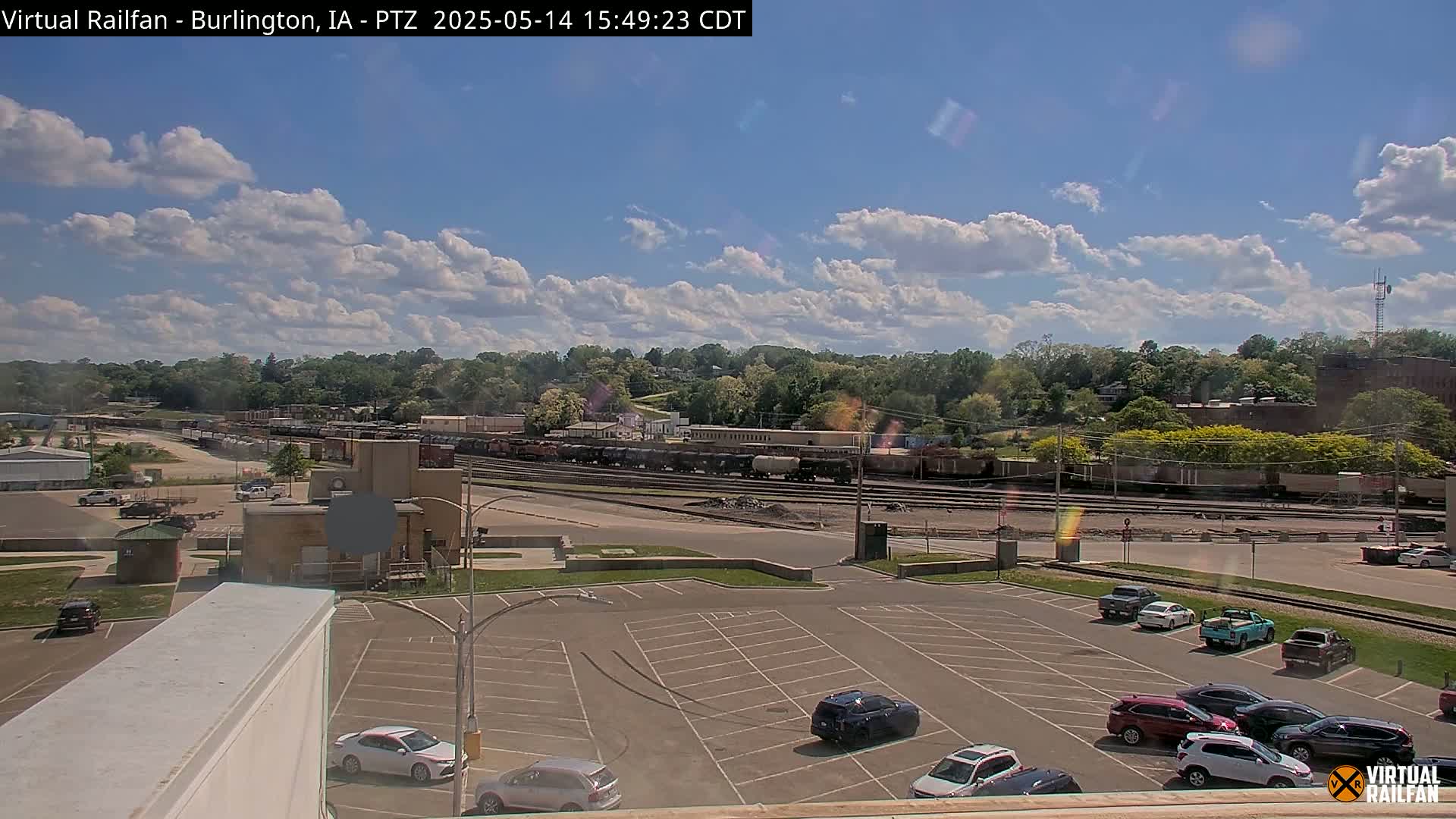 A partly cloudy day reveals a train yard with numerous rail cars, a parking lot with several vehicles, and a background of lush green trees.