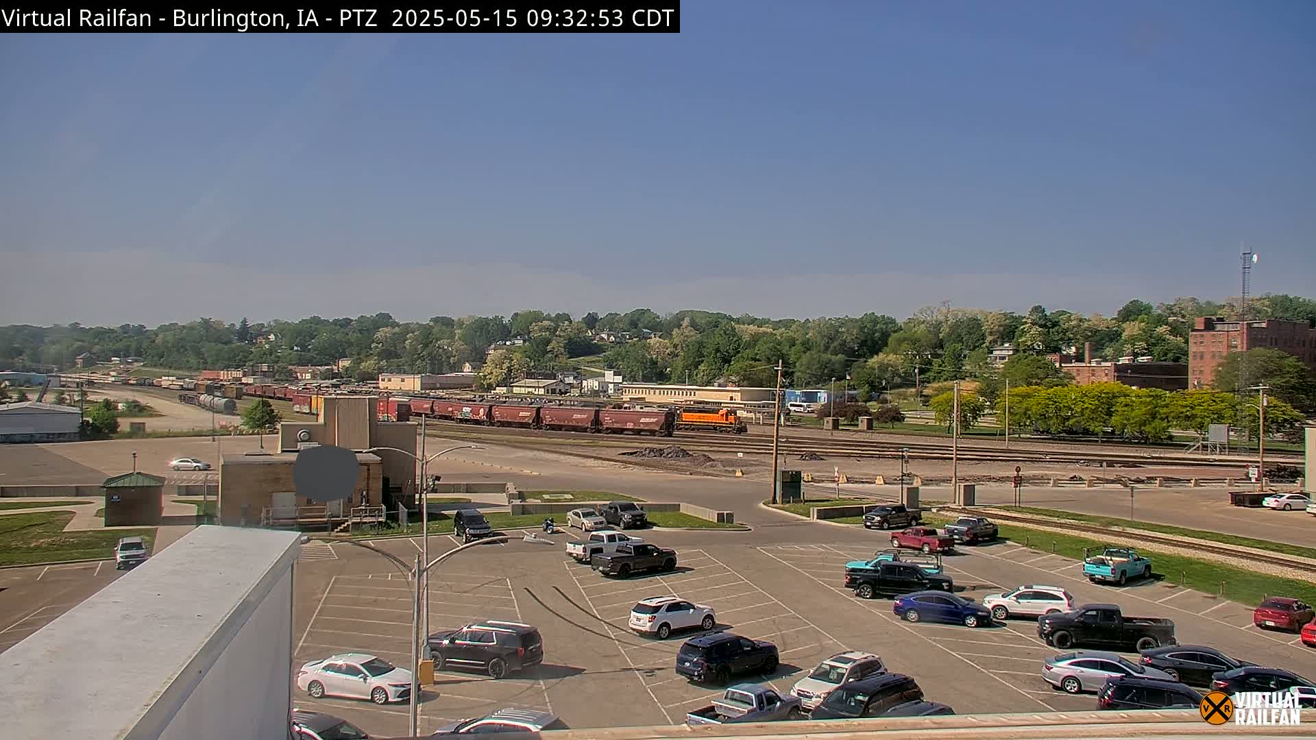 A long freight train is visible in the distance, under a clear blue sky, with a full parking lot of cars in the foreground.