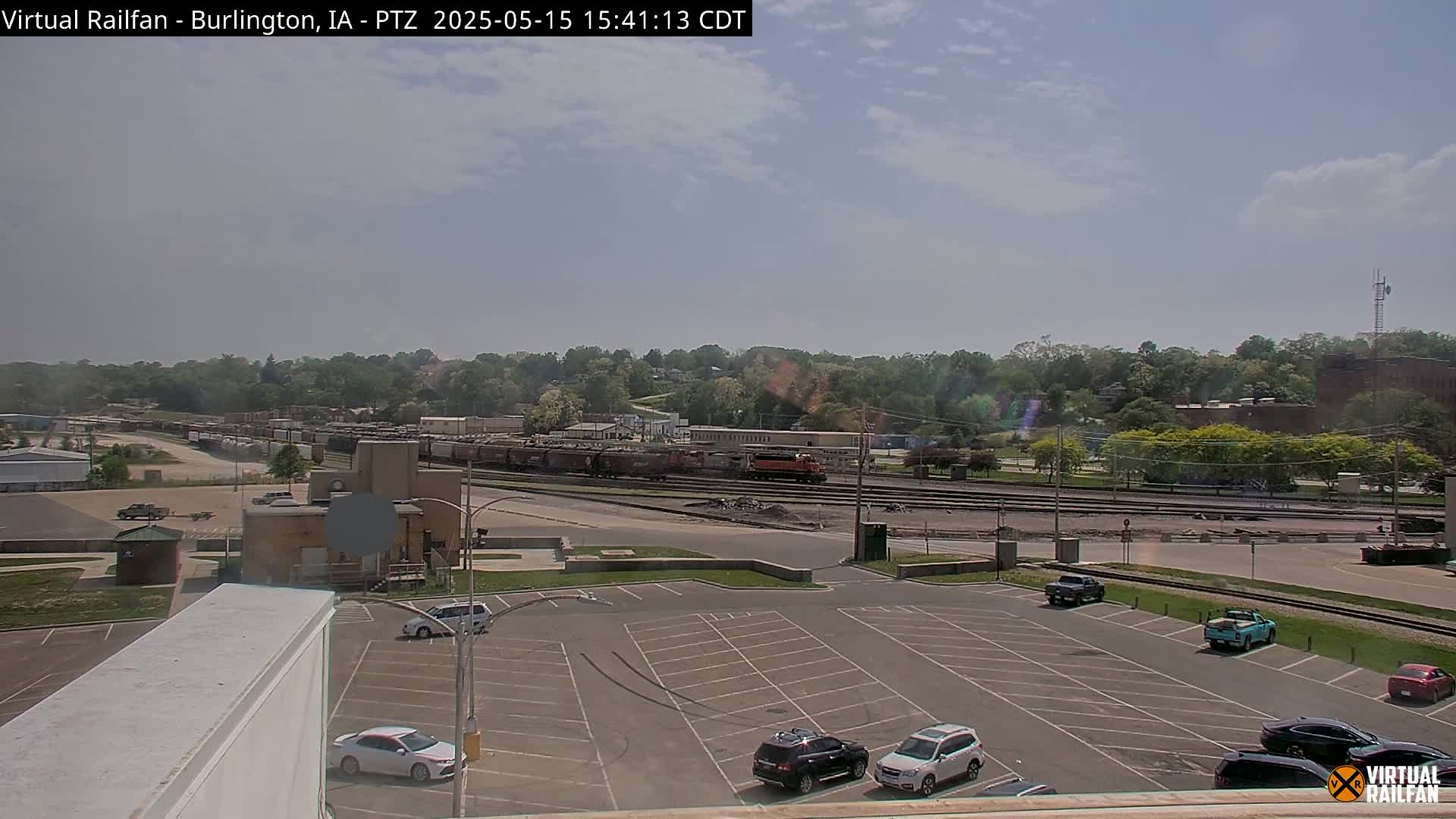 A mostly sunny day reveals a train yard with several rail cars, a parking lot with cars, and a tree-lined hill in the background.