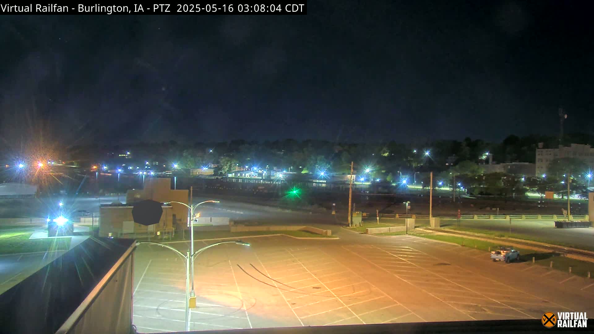 An empty, brightly lit parking lot at night overlooks a tree-lined area with buildings and streetlights in the distance under a clear, dark sky.