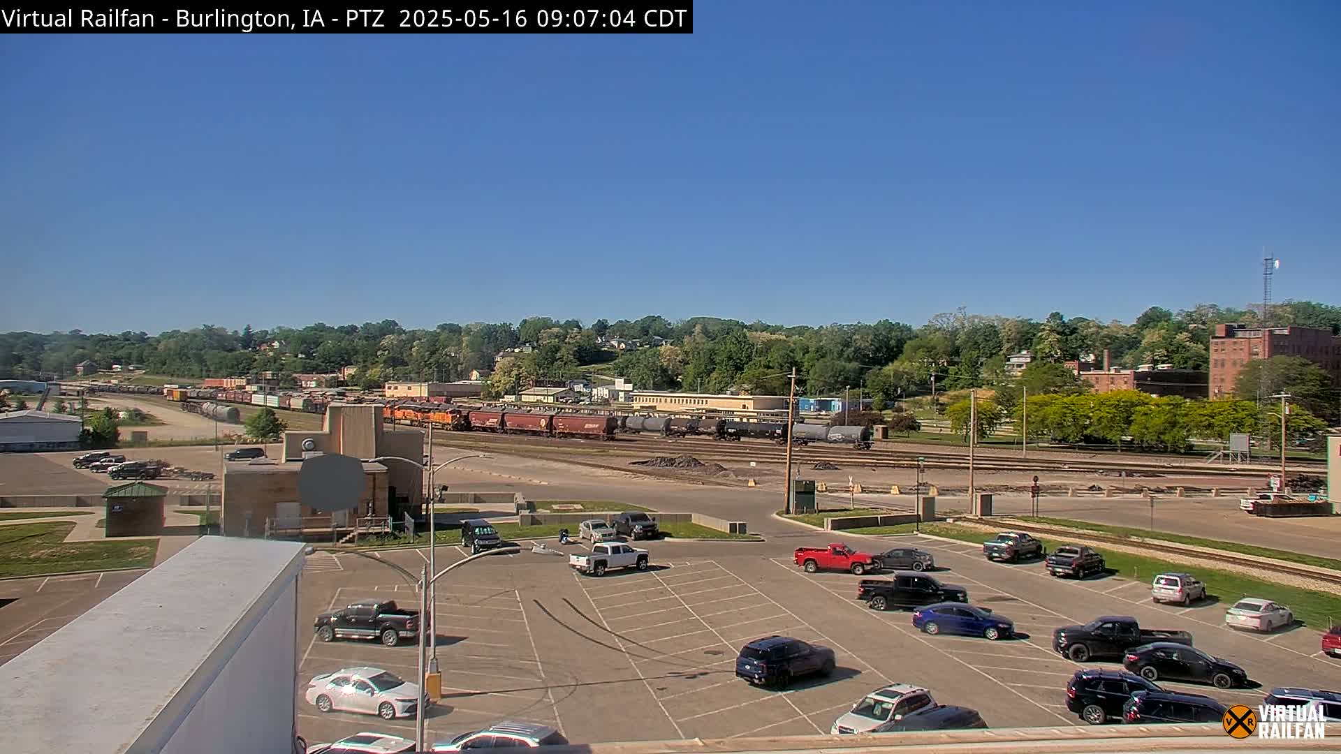 A long freight train sits in a rail yard next to a parking lot full of cars under a clear blue sky.