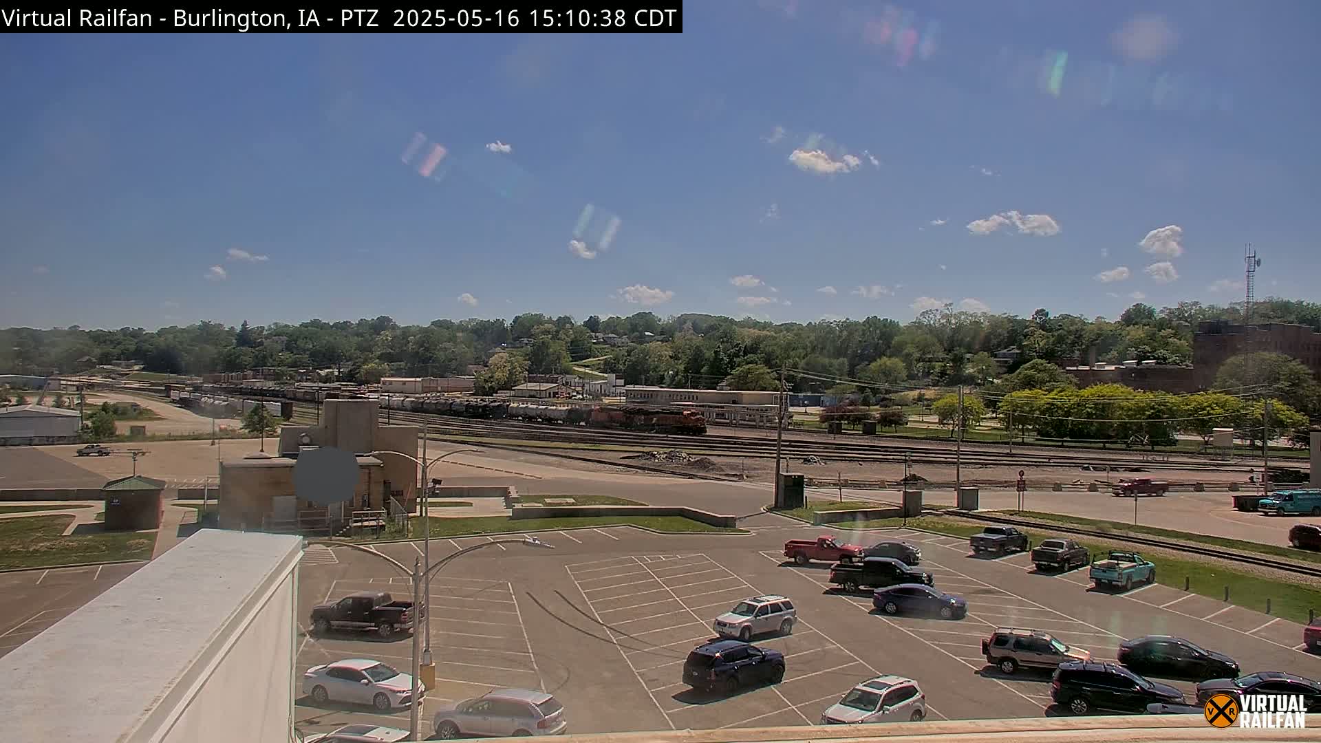 A mostly sunny day shows a train yard with a line of rail cars and several parked cars in a lot, all situated in front of a tree-lined hill.