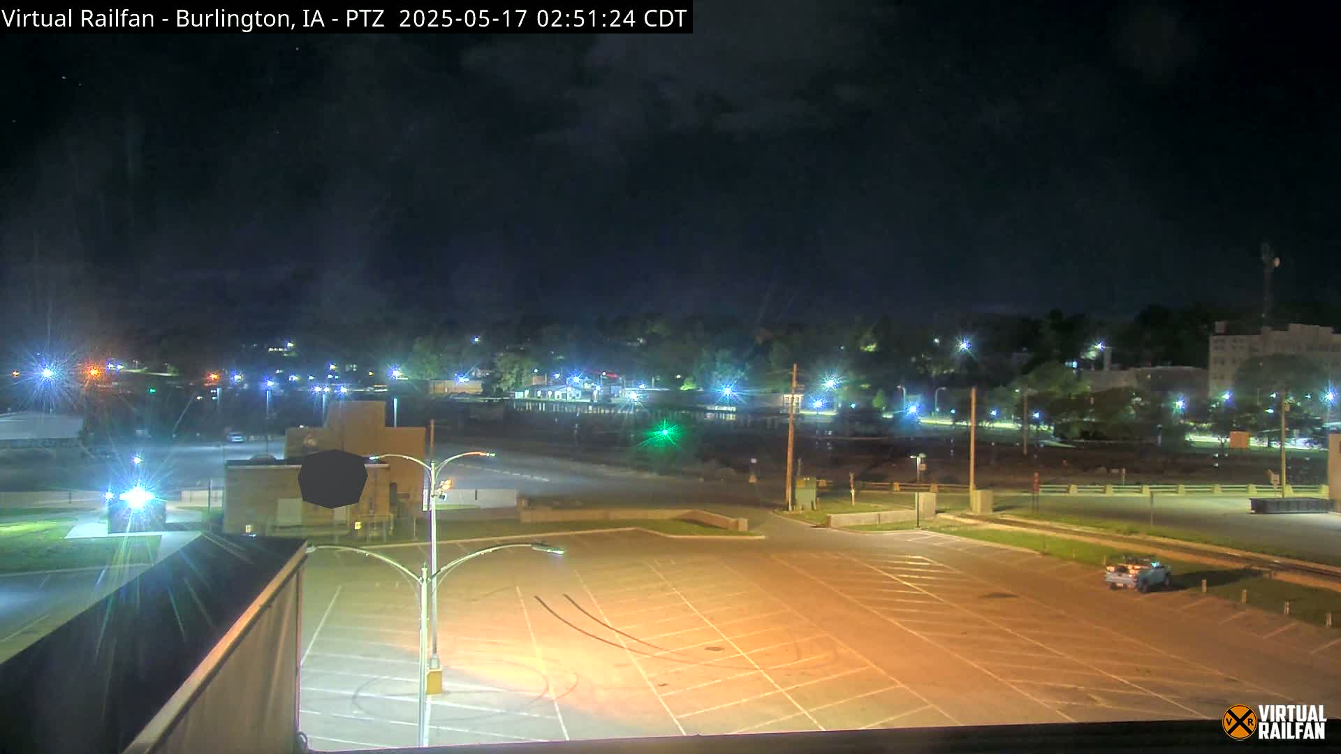 A mostly empty, illuminated parking lot at night, with a town’s lights visible in the background under a partly cloudy sky.