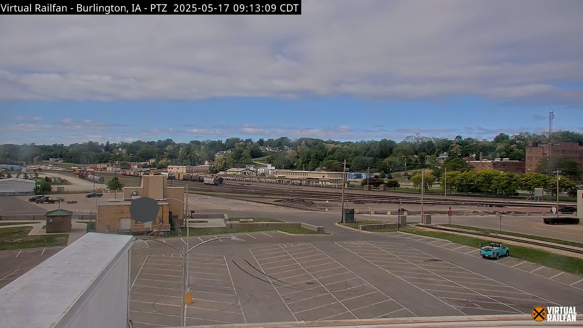 A mostly sunny day shows a train yard with several rail cars, a large empty parking lot, and a background of green trees and buildings.