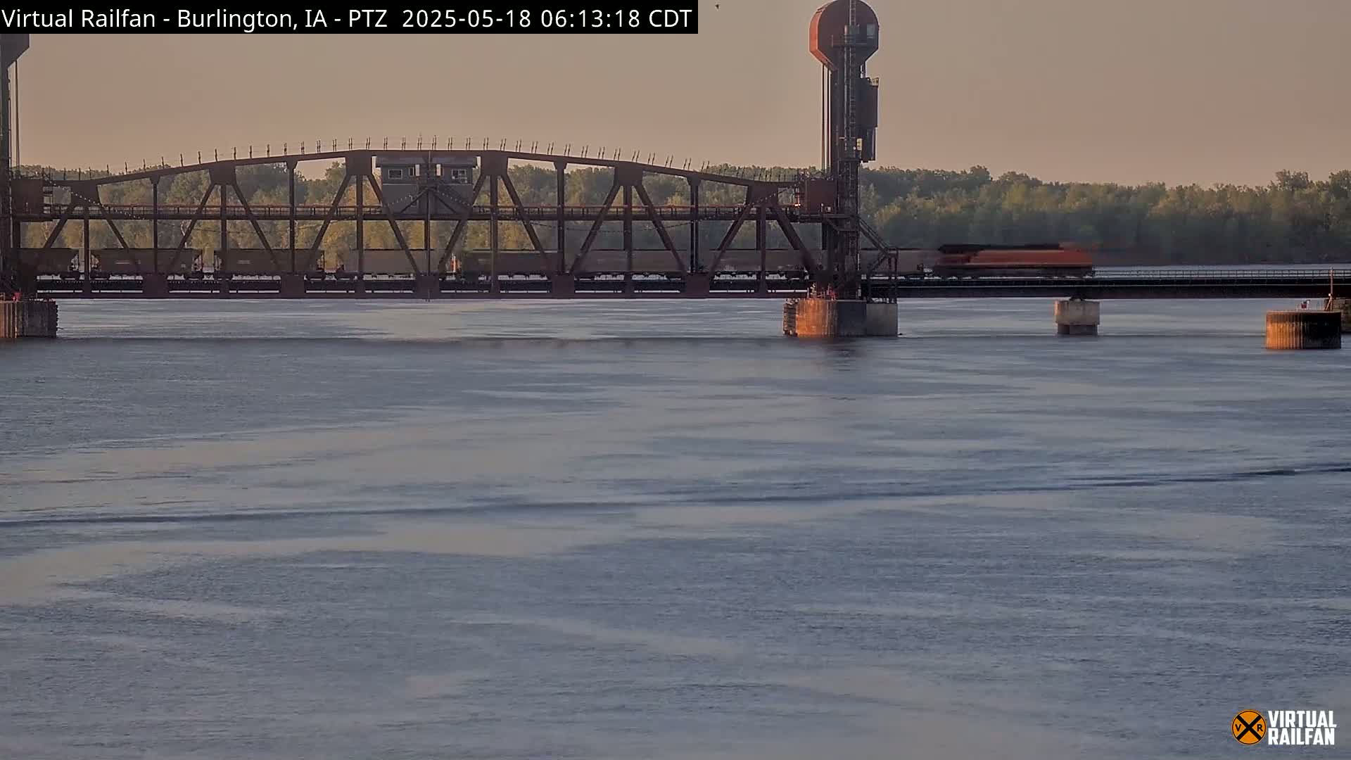 A train crosses a metal railroad bridge over a calm river under a twilight sky.