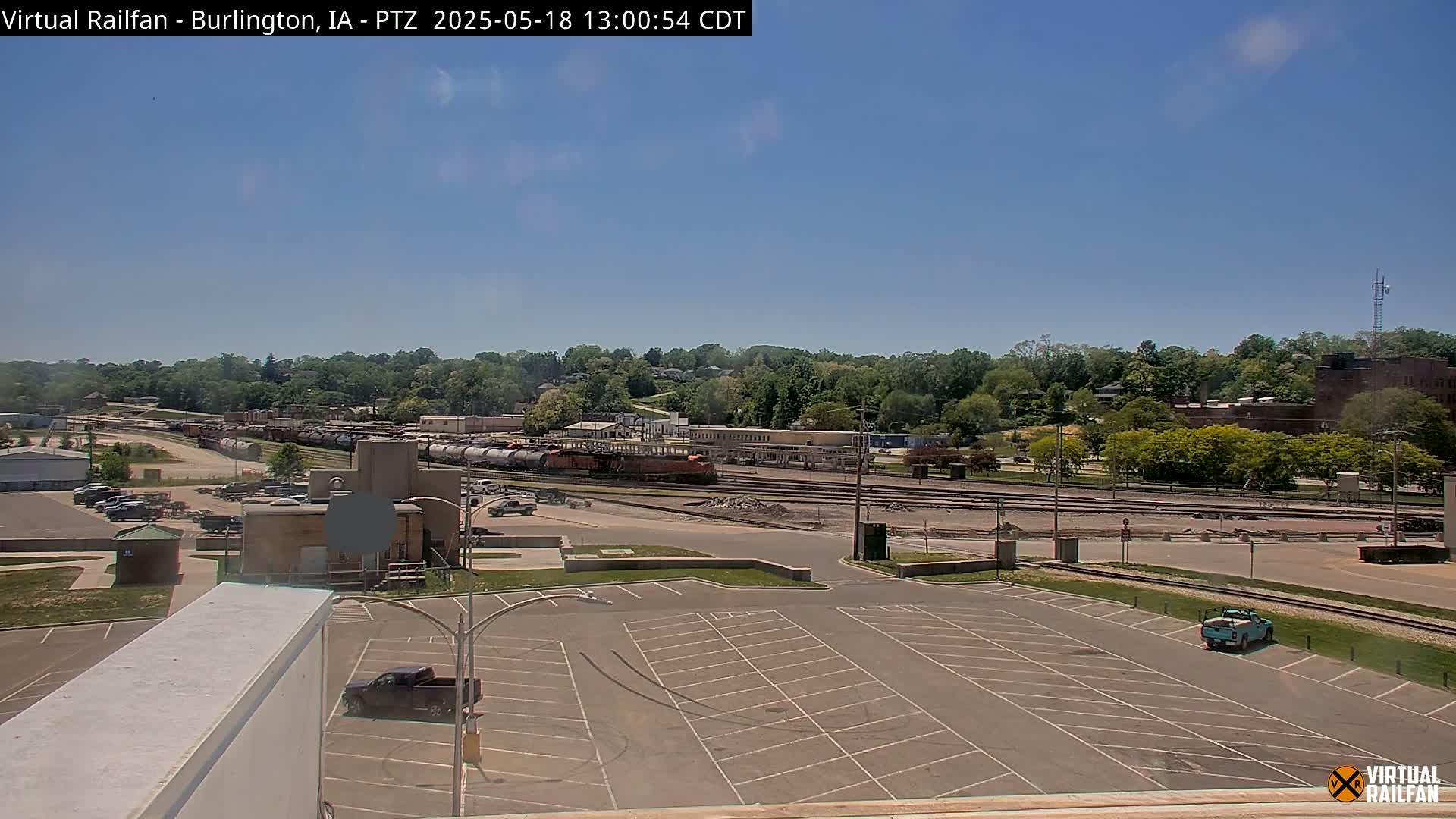 A mostly sunny day reveals a large freight train in a rail yard, adjacent to a parking lot and a tree-lined hill.