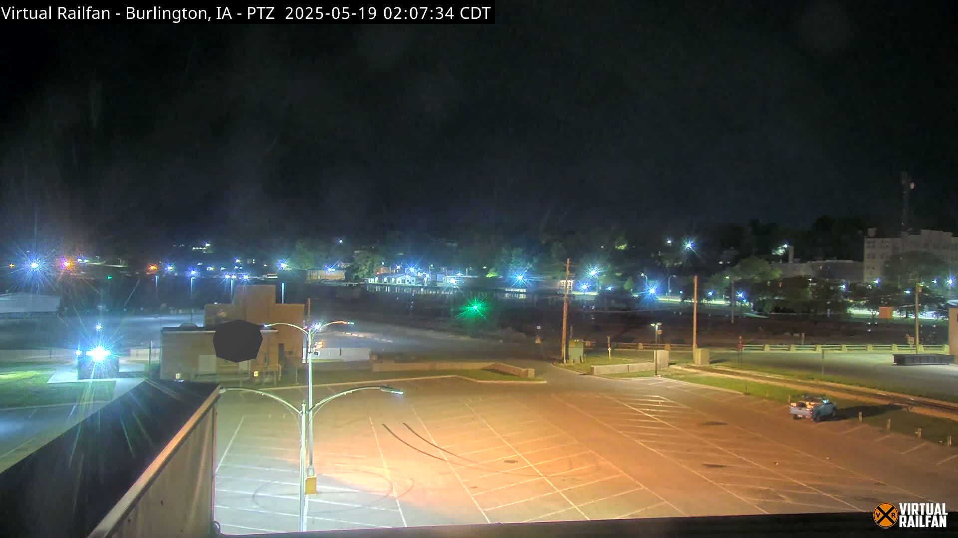 An empty, illuminated parking lot at night overlooks a dimly lit town with trees and buildings under a clear, dark sky.