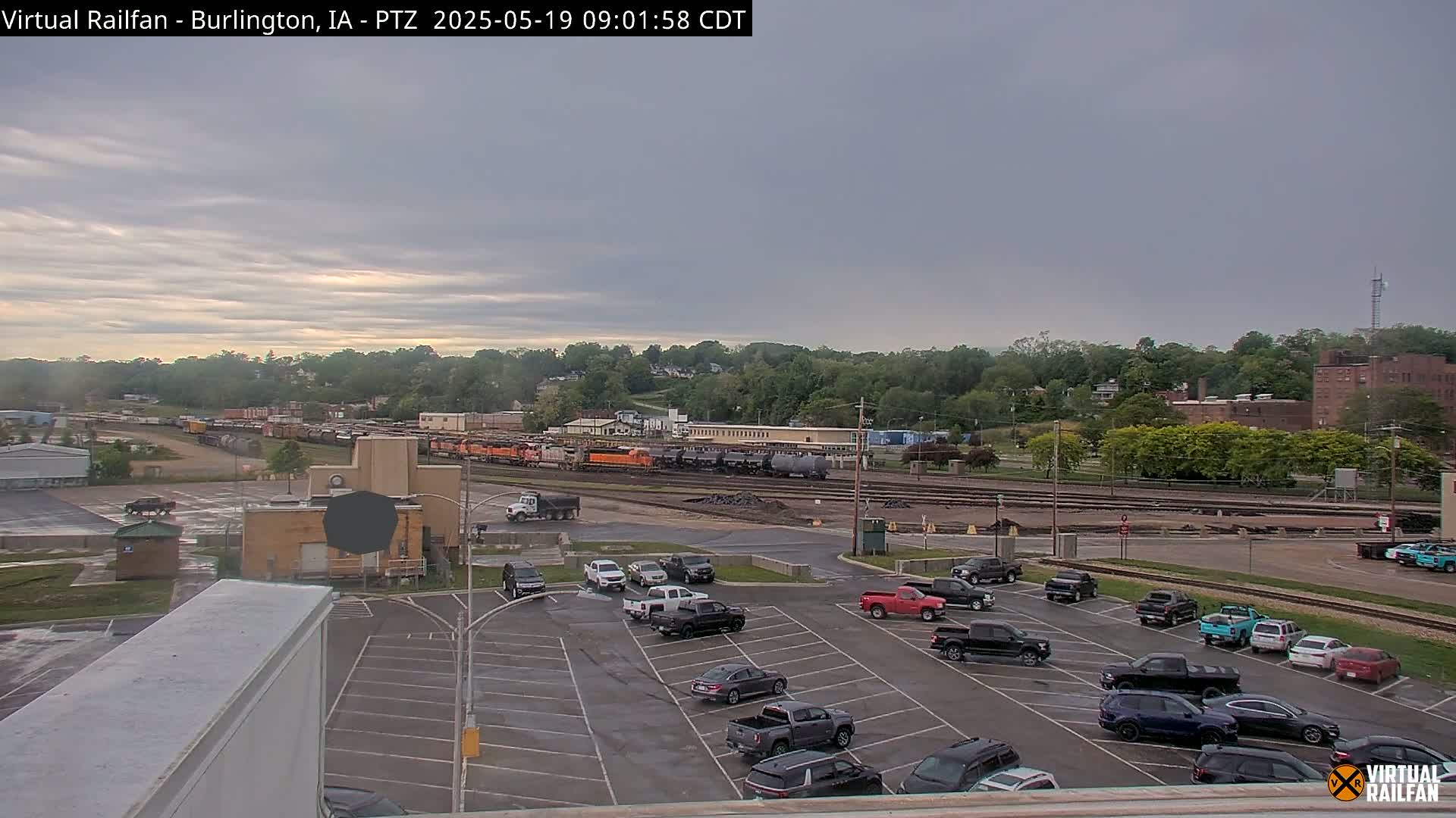 A mostly cloudy day reveals a train yard with several cars parked in a lot, surrounded by industrial buildings and green trees.