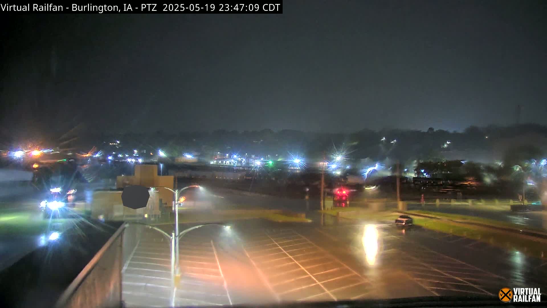 A nighttime, rainy scene shows a wet parking lot and roadway with distant lights and vehicles under a dark sky.
