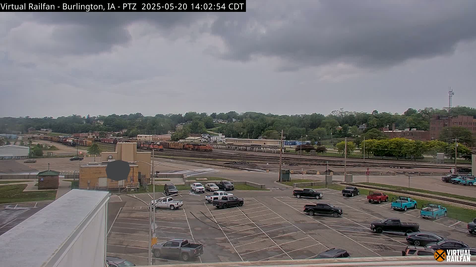 A mostly cloudy day reveals a freight train and several cars parked in a lot, with a line of trees and buildings in the background.