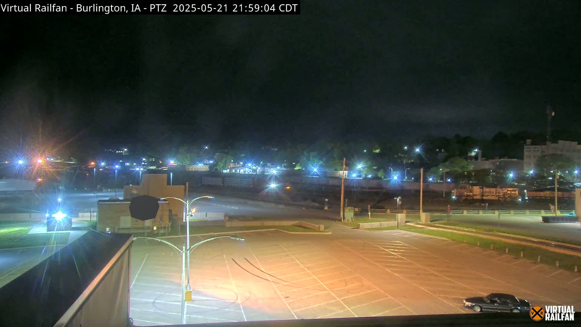 A nighttime view of a mostly empty parking lot next to railroad tracks with a train visible in the distance under a clear, dark sky.
