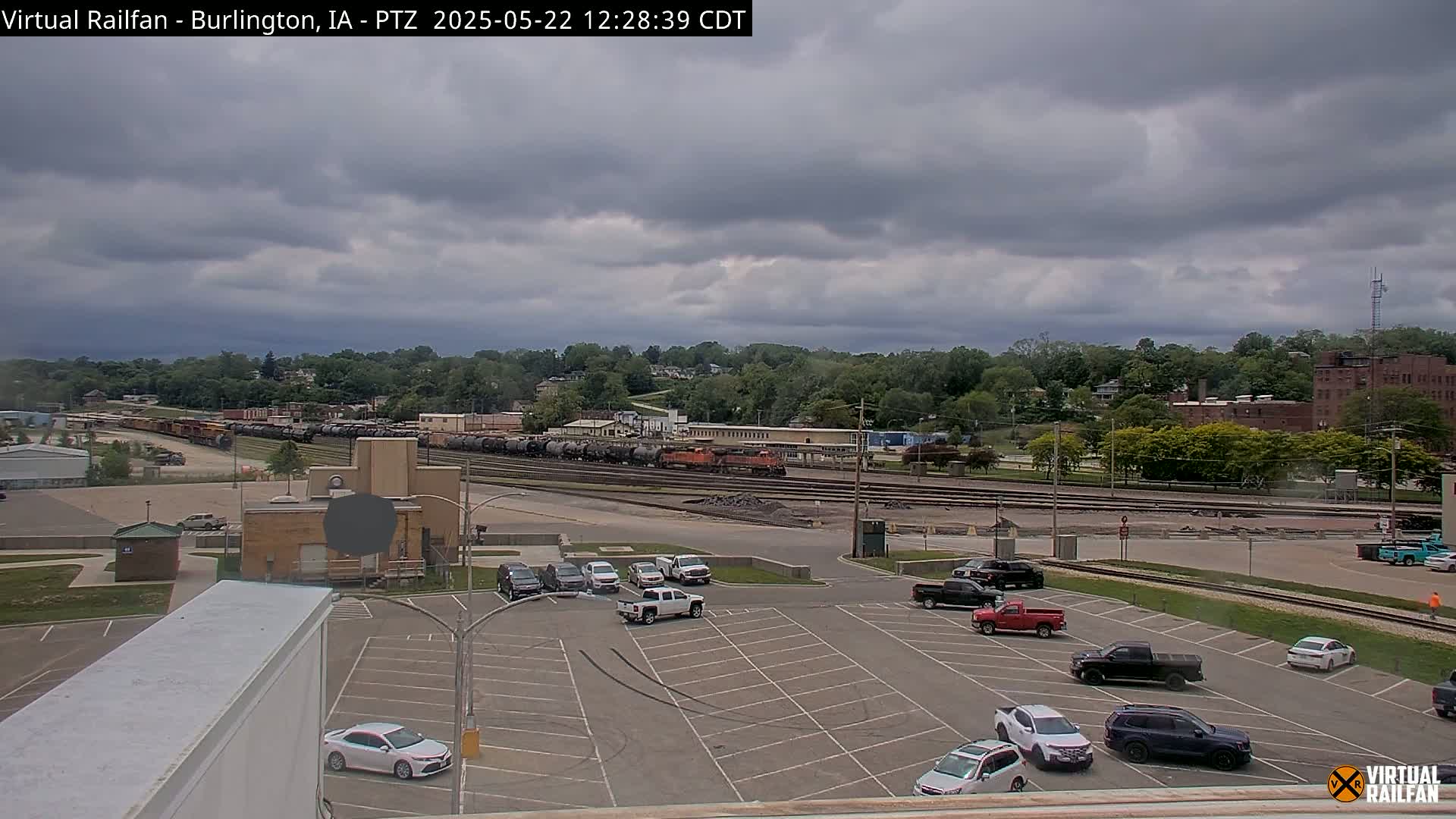 A mostly cloudy day reveals a train yard with several railcars, a parking lot with cars, and a background of trees and buildings.