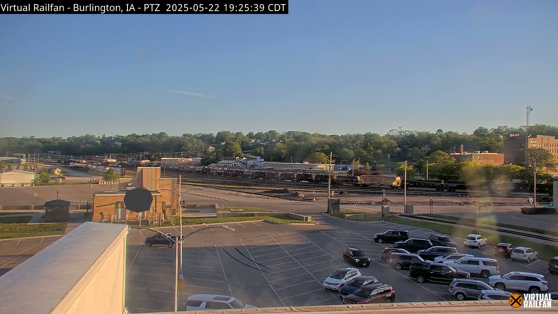 A long train sits in a rail yard adjacent to a large parking lot filled with cars, all under a clear, sunny sky.