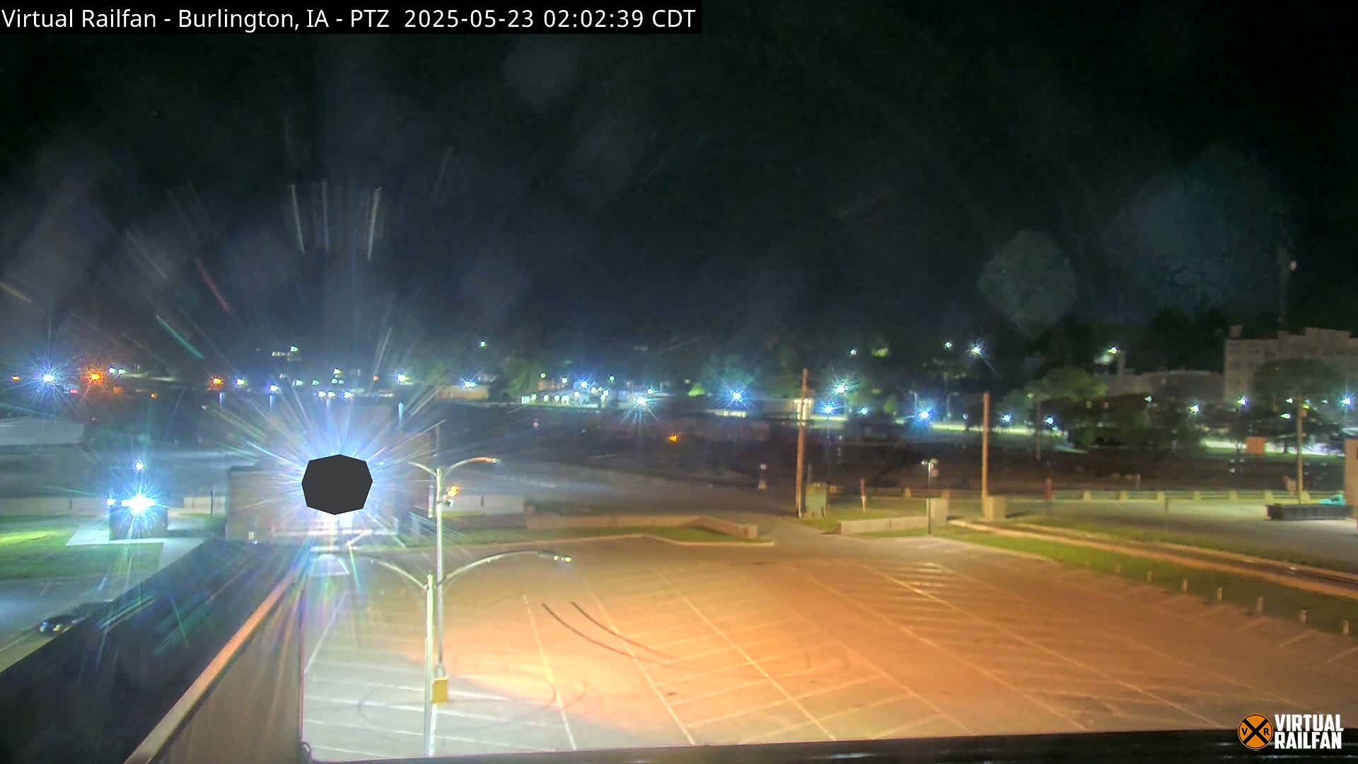 A nighttime, outdoor view of a mostly empty parking lot with streetlights illuminating a surrounding area with buildings and trees.