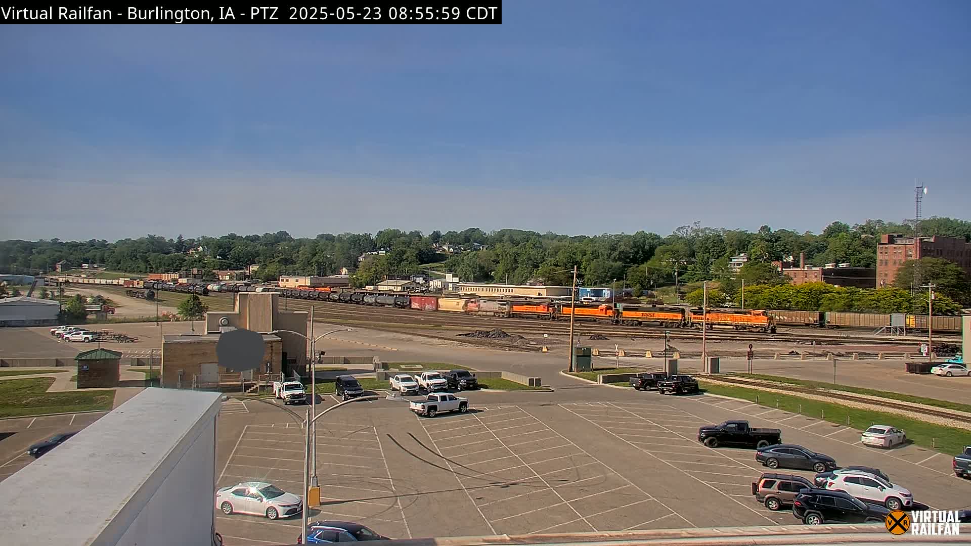 A long freight train sits in a rail yard under a clear blue sky, with several cars parked in a lot in the foreground.