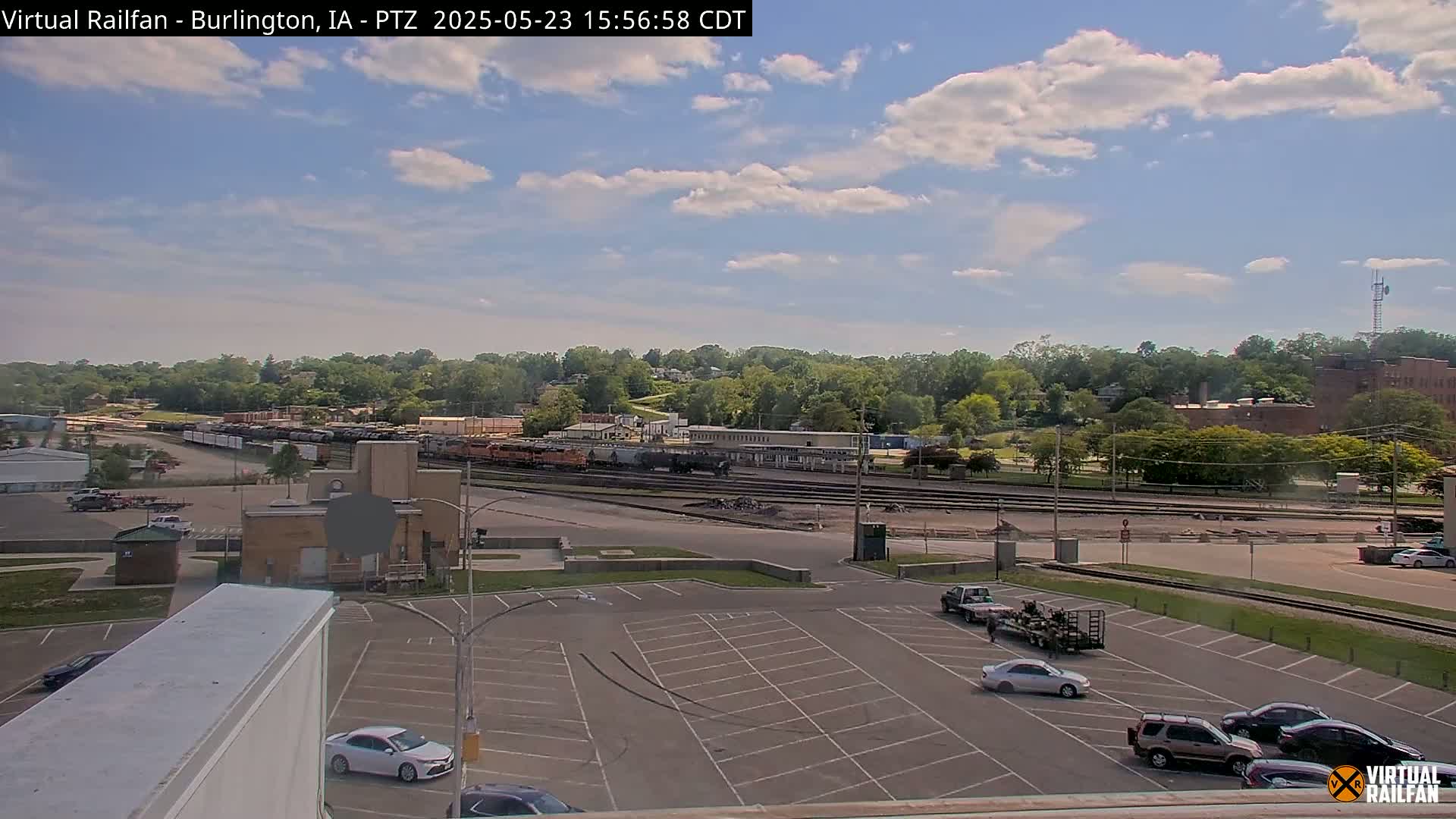 A mostly sunny day shows a railway yard with several train cars, a parking lot with cars, and a background of green trees and buildings.