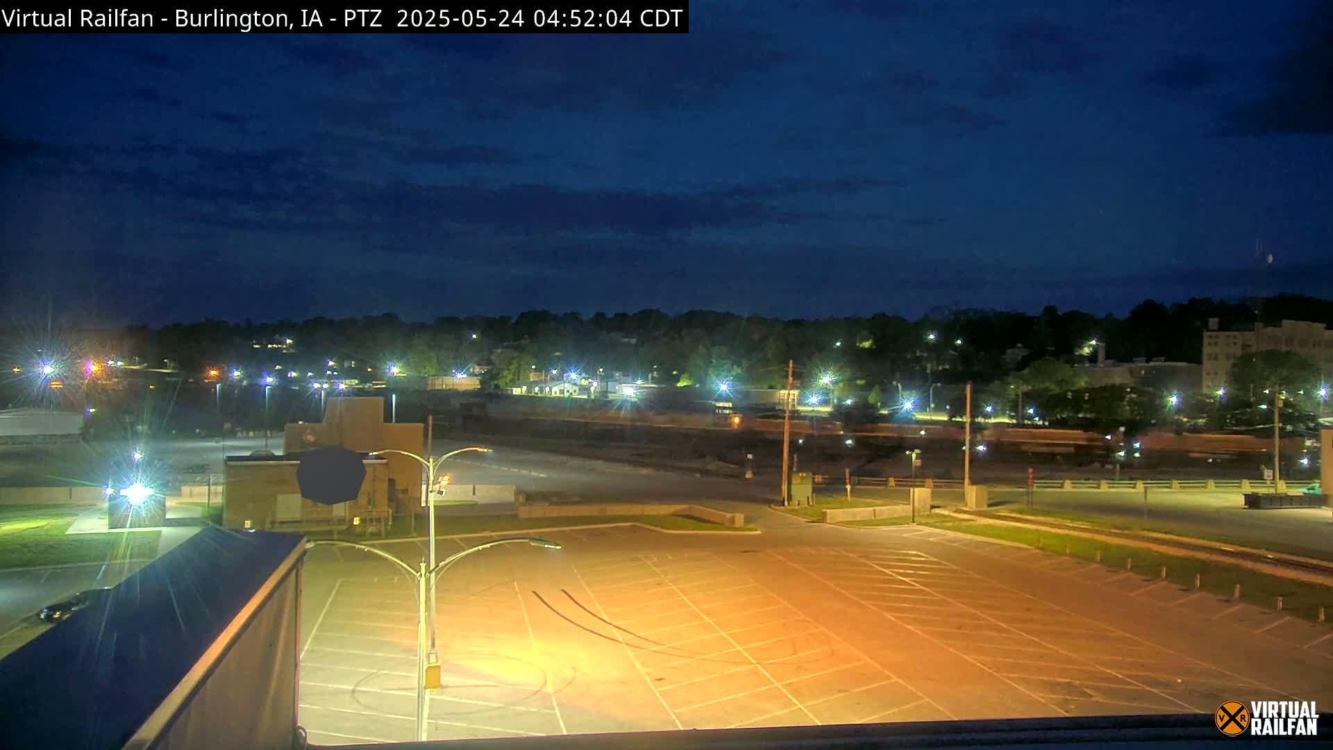 An empty, illuminated parking lot overlooks a dark, tree-lined area with distant lights and railway tracks under a twilight sky.