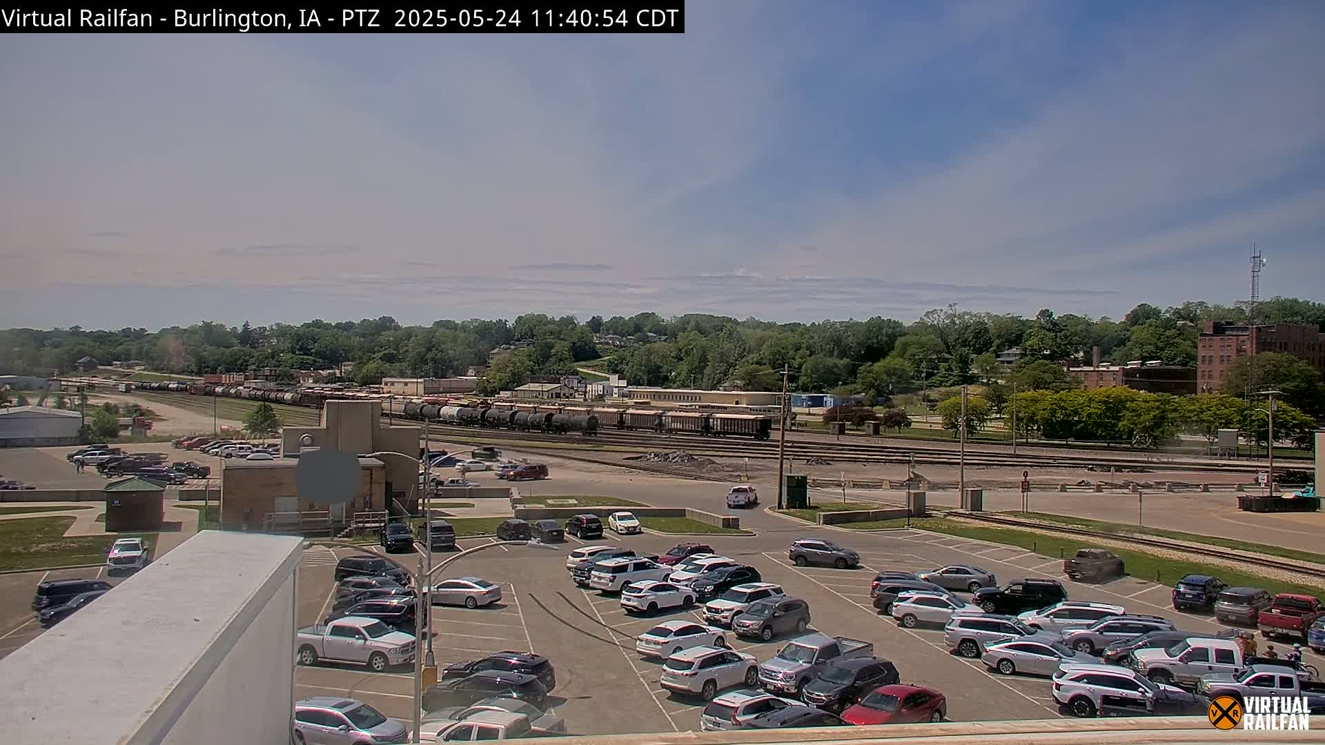 A long freight train sits in a rail yard next to a large parking lot filled with cars, under a mostly sunny sky.