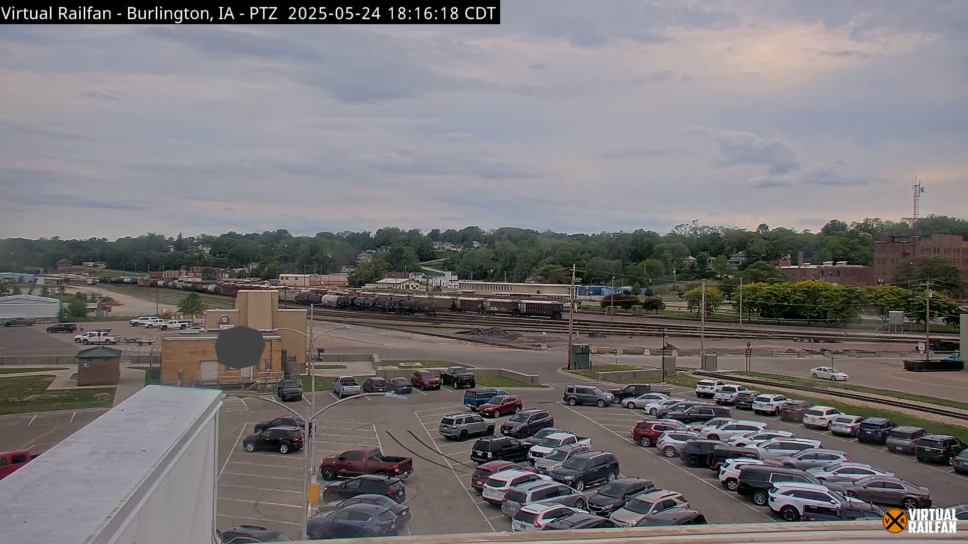 A mostly cloudy day reveals a large train yard with numerous rail cars, adjacent to a sizable parking lot filled with cars, all situated in front of a tree-lined hill with buildings in the distance.