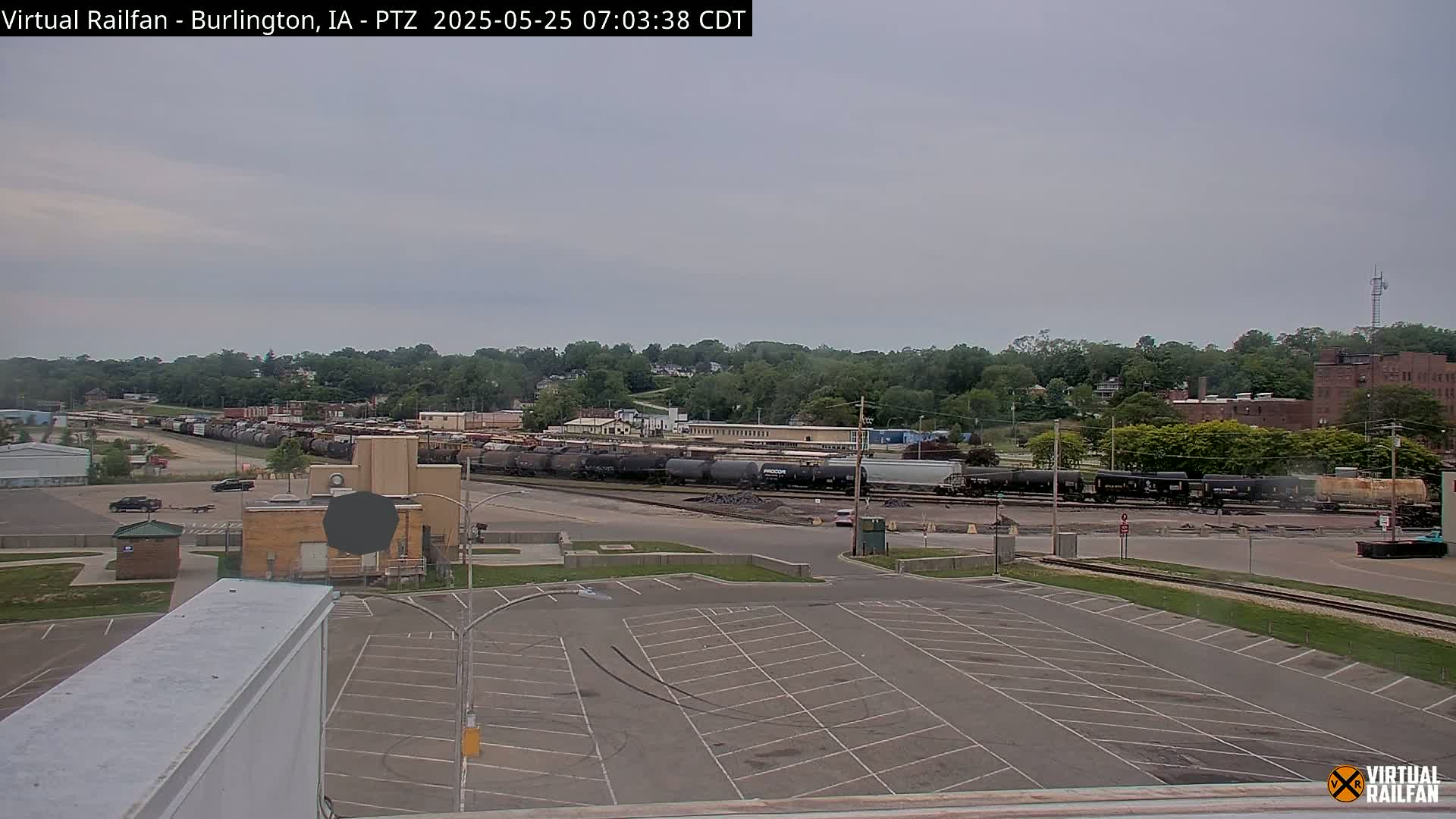 A large empty parking lot overlooks a train yard filled with numerous railcars under an overcast sky.