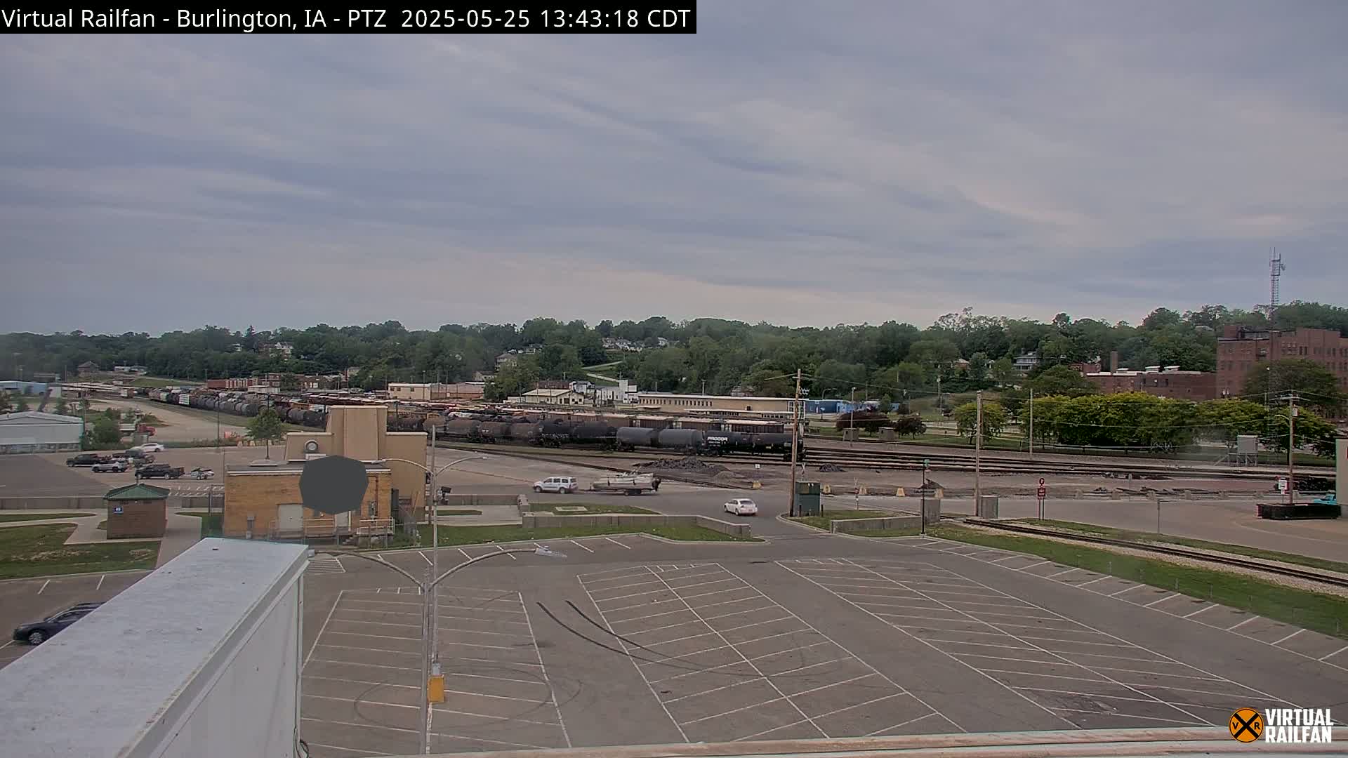 A large empty parking lot overlooks a train yard filled with numerous rail cars under a cloudy sky.