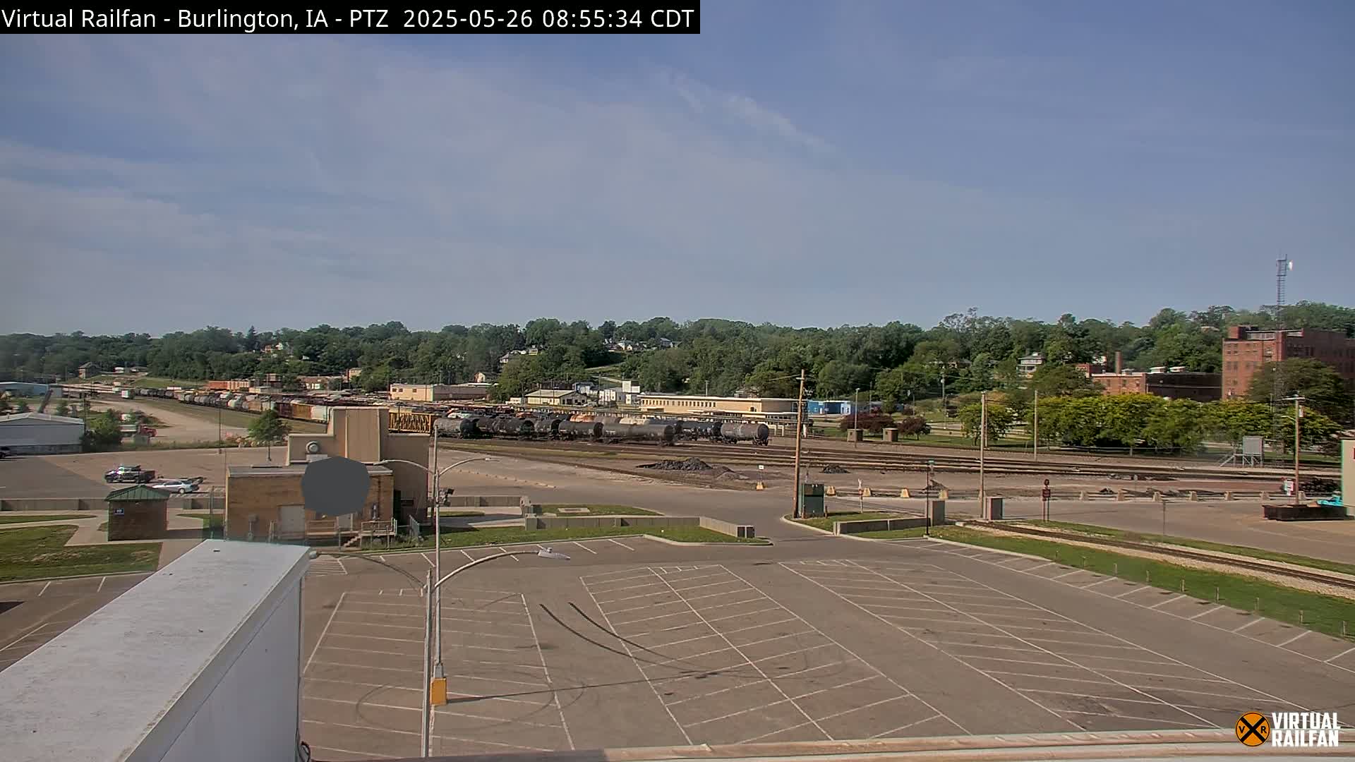 An empty parking lot overlooks a rail yard with numerous train cars under a mostly clear, sunny sky.