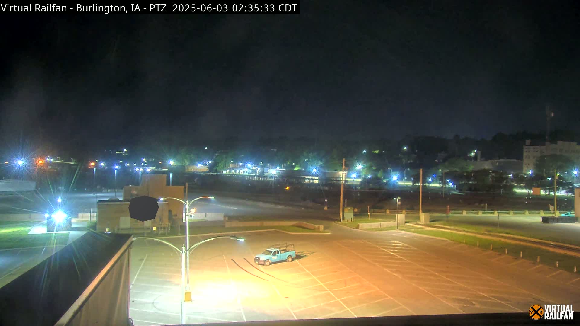 A mostly empty parking lot at night is illuminated by streetlights, with a single blue truck parked in the foreground and a distant town faintly visible under a dark sky.