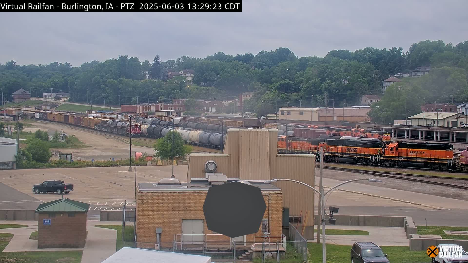 A long freight train passes by a rail yard under an overcast sky, with several buildings and a pickup truck visible in the foreground.
