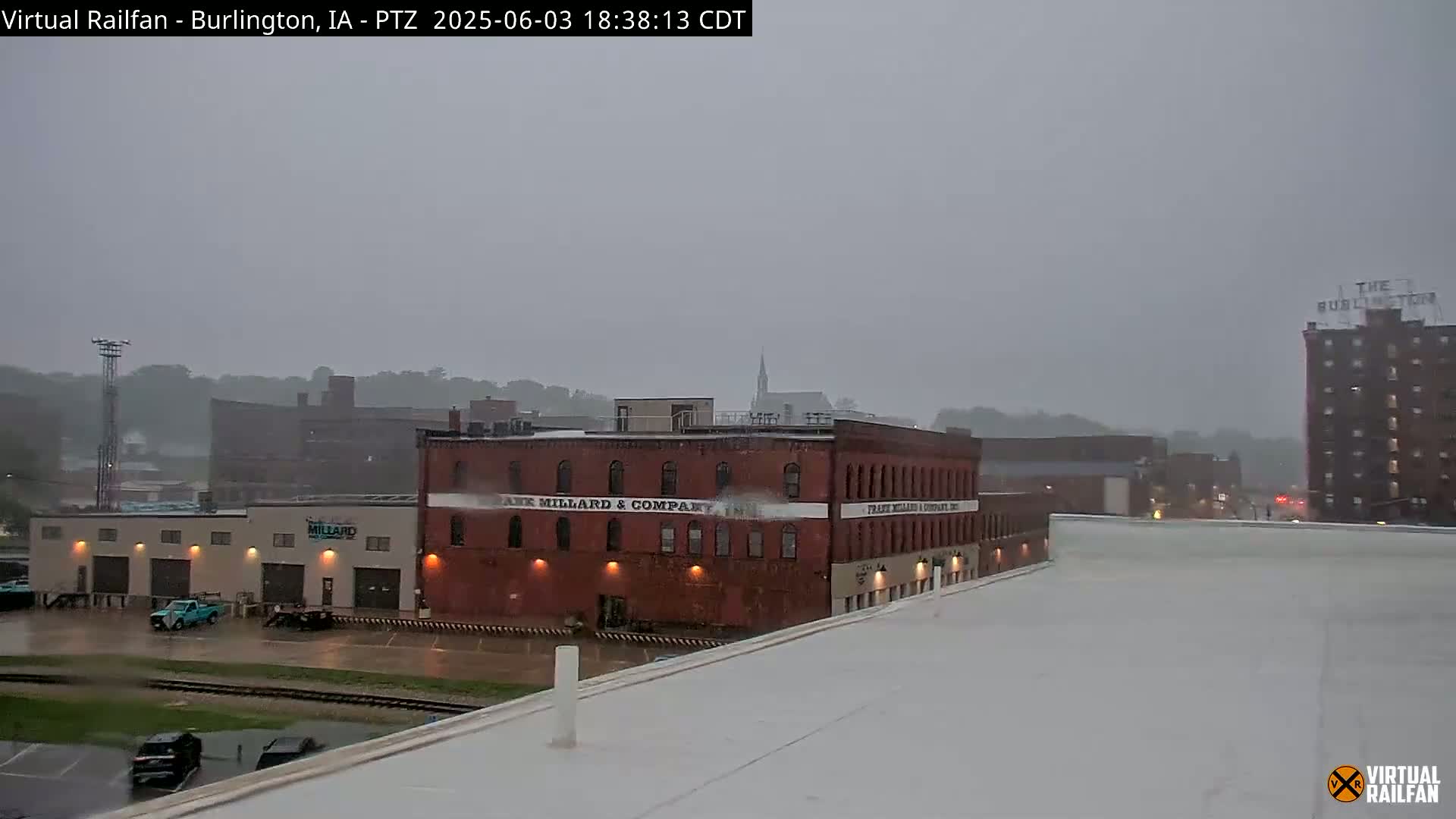 A rainy view of a city block featuring several brick buildings, some with lights on, seen from a rooftop under a gray sky.