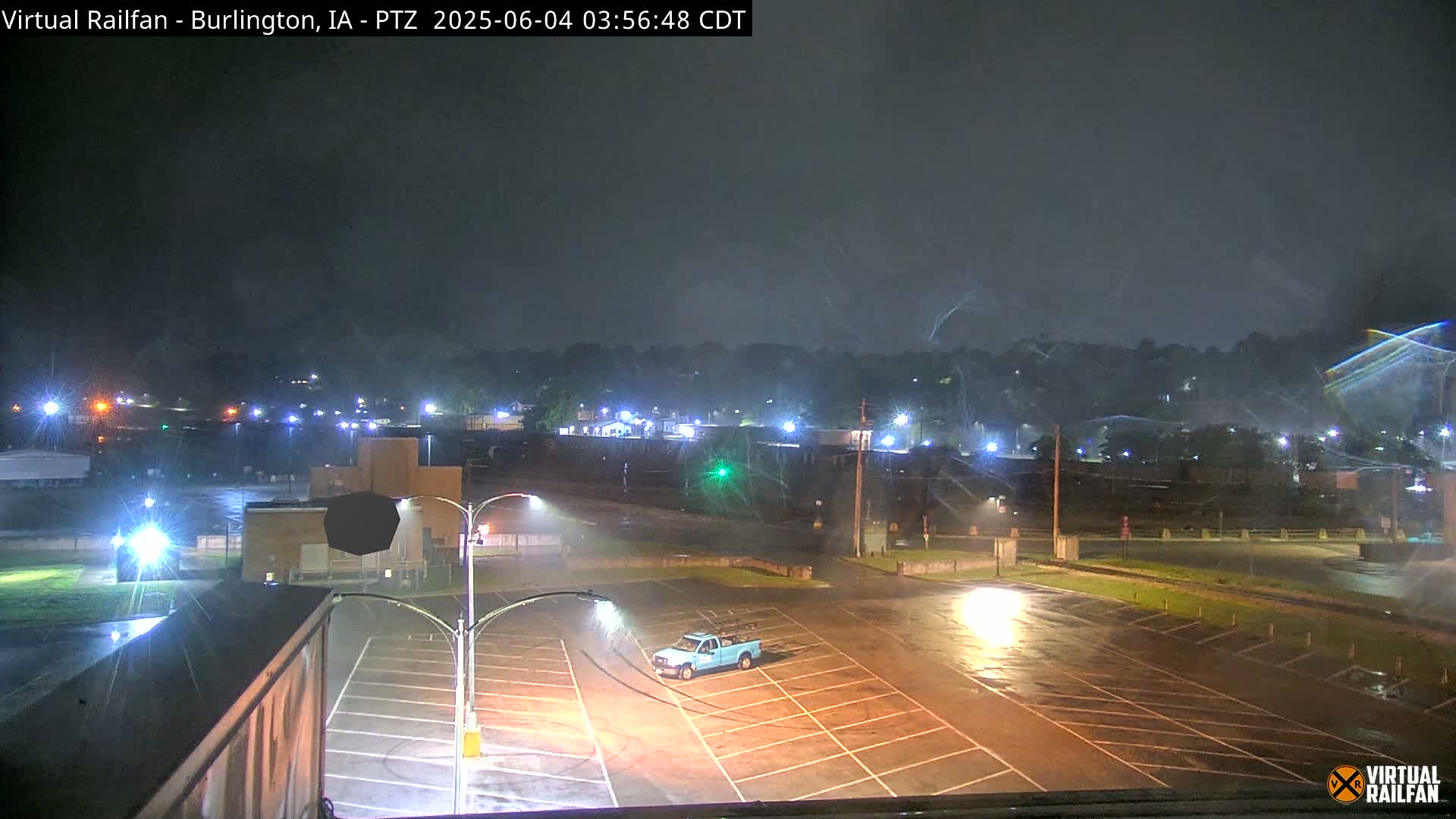 A rain-slicked empty parking lot at night, under a dark sky with streaks of rain, is illuminated by streetlights and shows a single light blue pickup truck parked in the lot, with a distant view of buildings and trees.