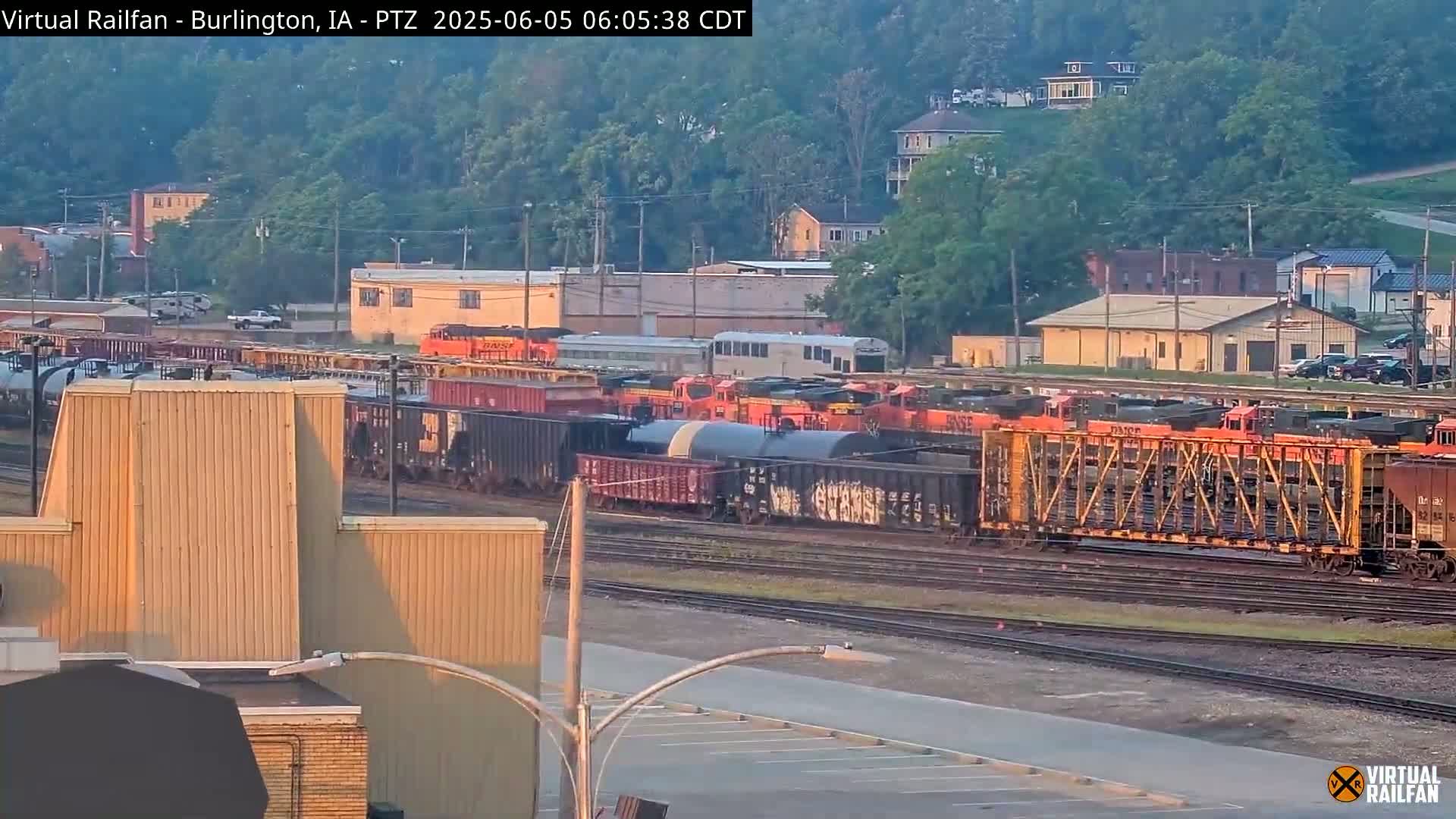 A long freight train sits on tracks in a rail yard at sunrise, with buildings and a hill visible in the background.