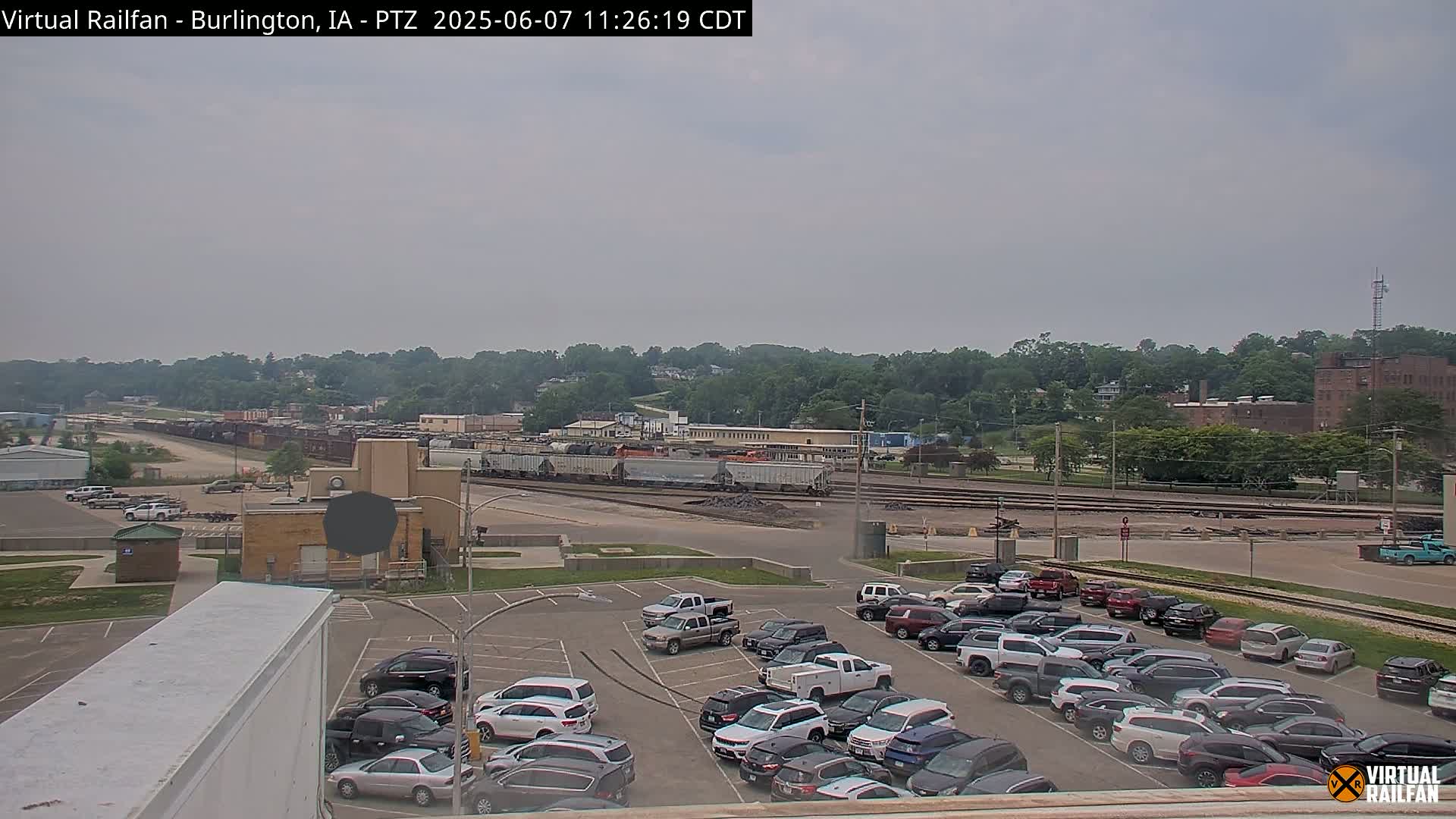 A large parking lot filled with cars is situated in front of a railway yard containing numerous train cars, all under an overcast sky.