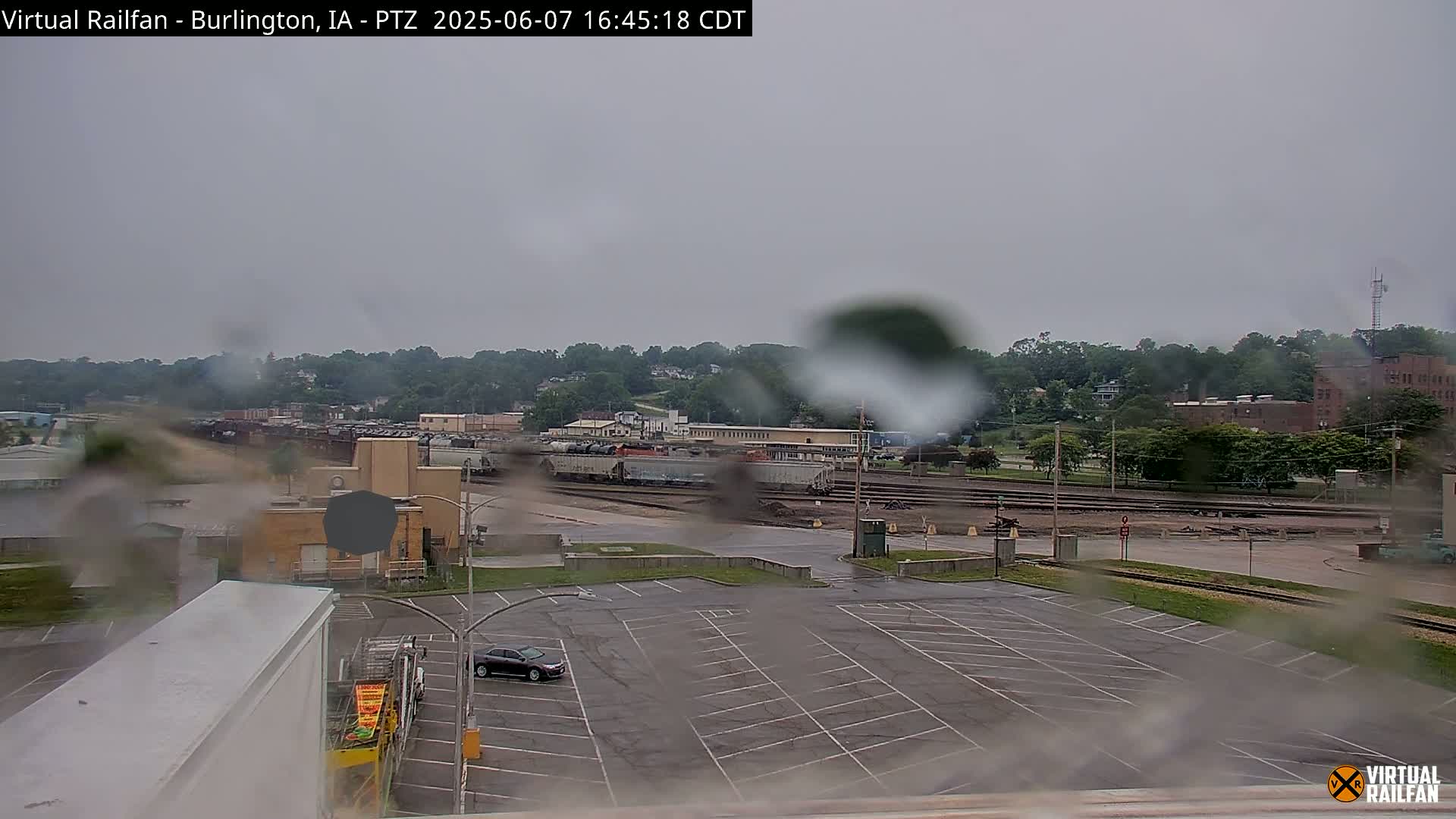 A rainy view of a railroad yard with several train cars, a mostly empty parking lot, and a line of buildings in the background.