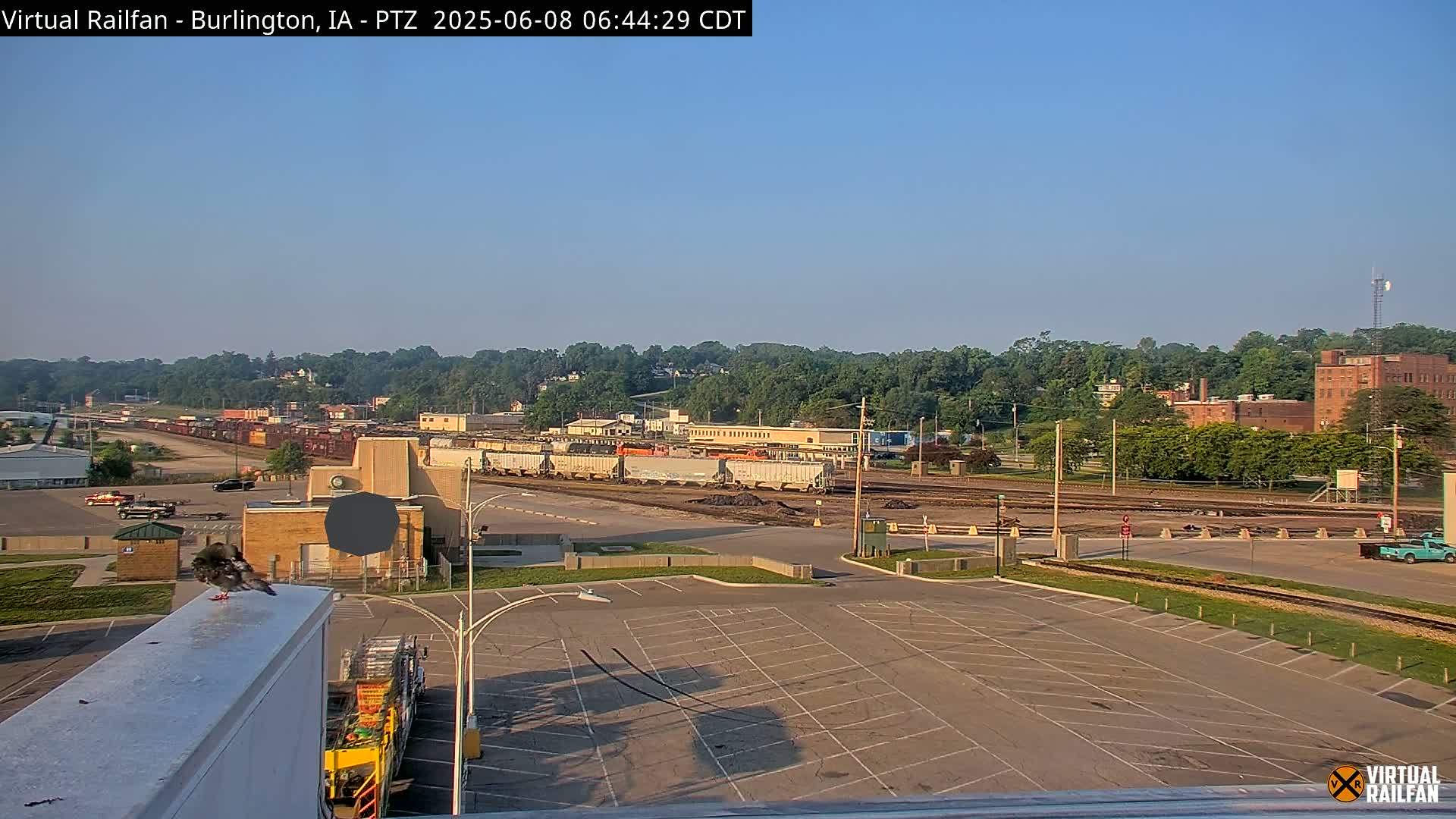 A large empty parking lot overlooks a train yard with several rail cars, a few buildings, and a tree-lined hill in the background under a clear, sunny sky.