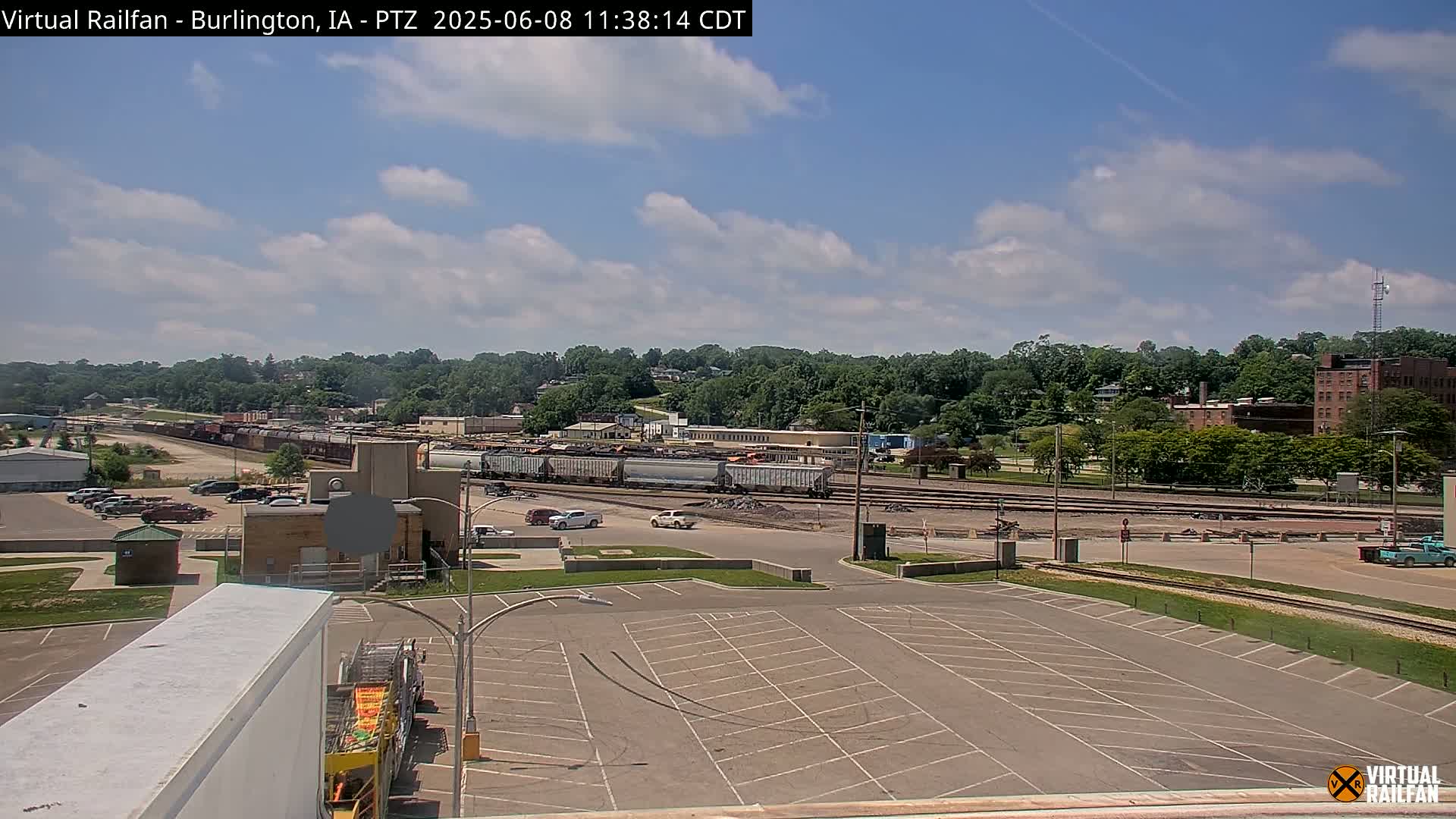 A partly cloudy day reveals a train yard with several railcars, adjacent parking lots, and a tree-lined hillside in the background.