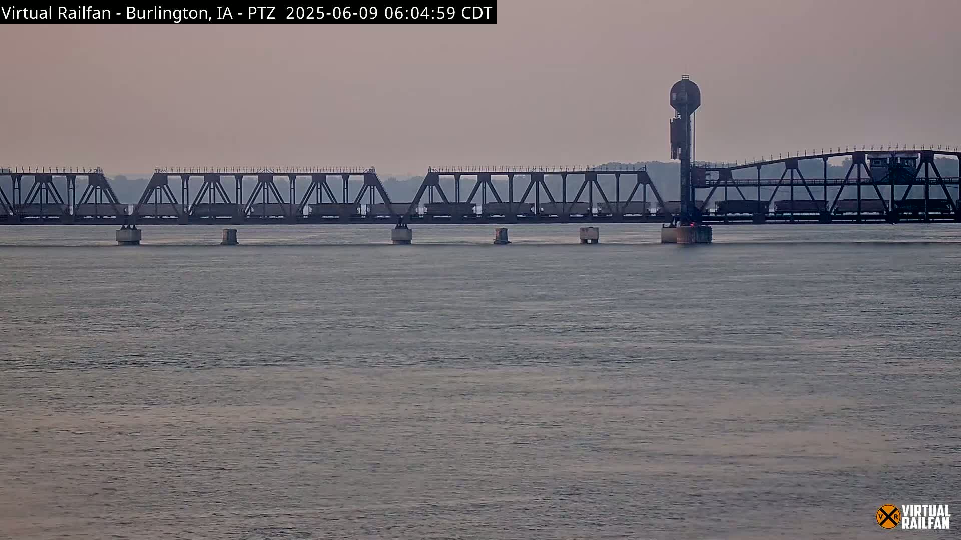 A long freight train crosses a metal truss bridge over calm water under a hazy sky.