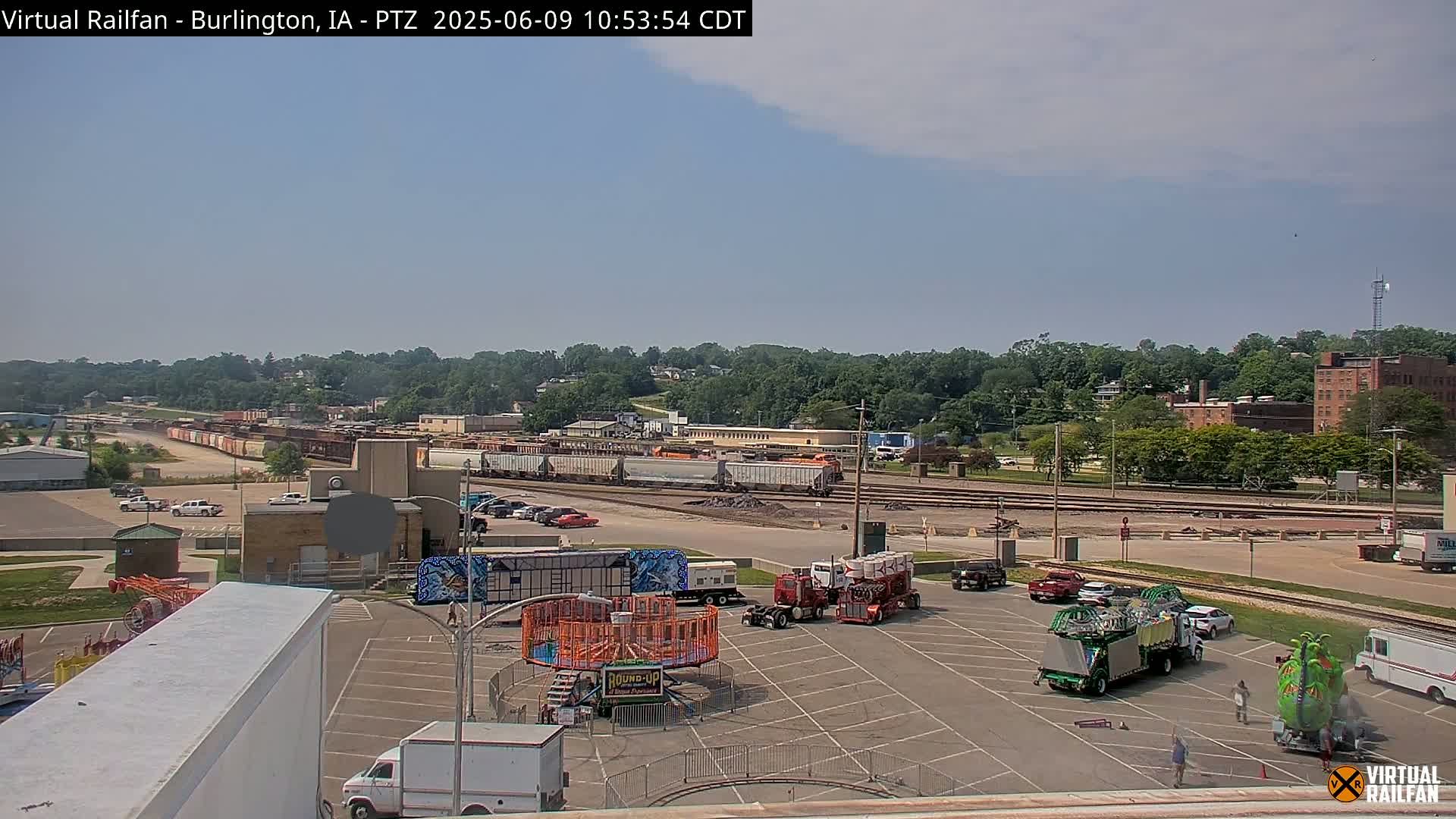 A mostly sunny day shows a train yard with numerous rail cars, a carnival midway set up in a parking lot, and a tree-lined cityscape in the background.