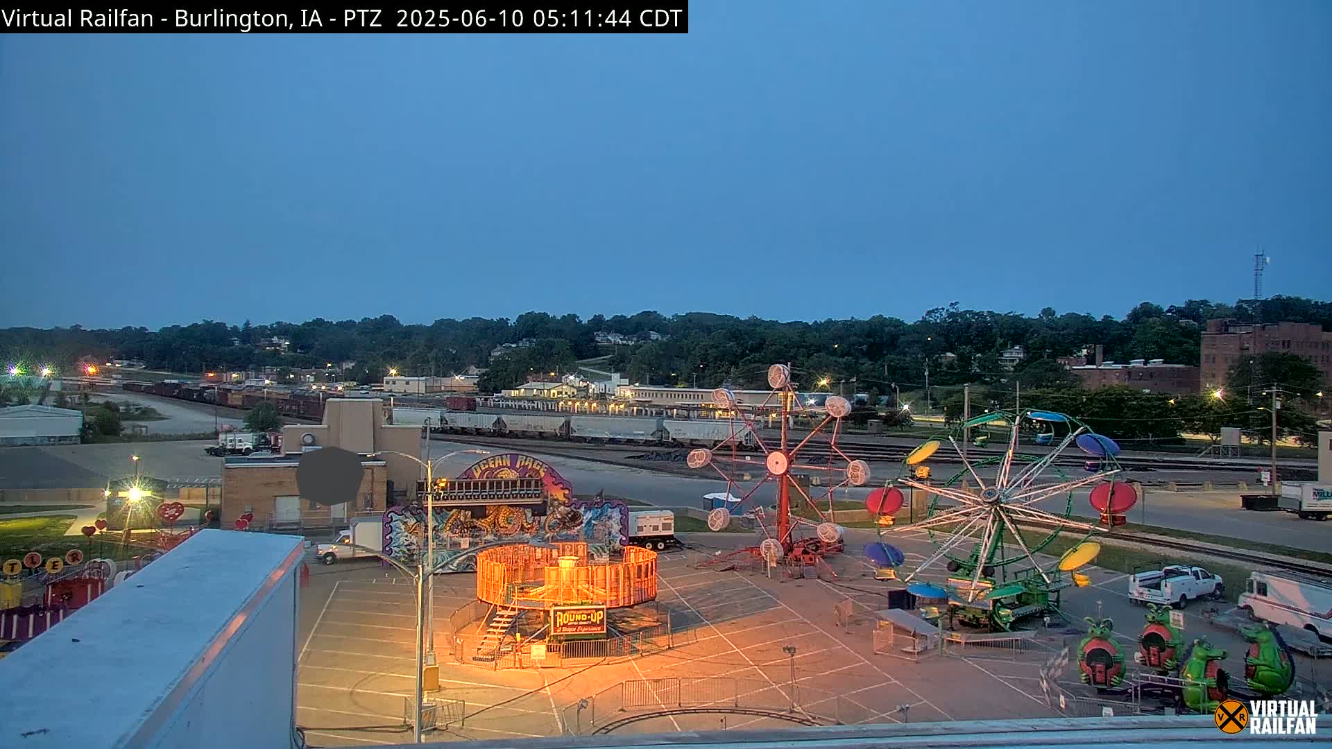 A nighttime view of a carnival midway with several rides set up in a parking lot, next to railroad tracks and a town situated on a hill.