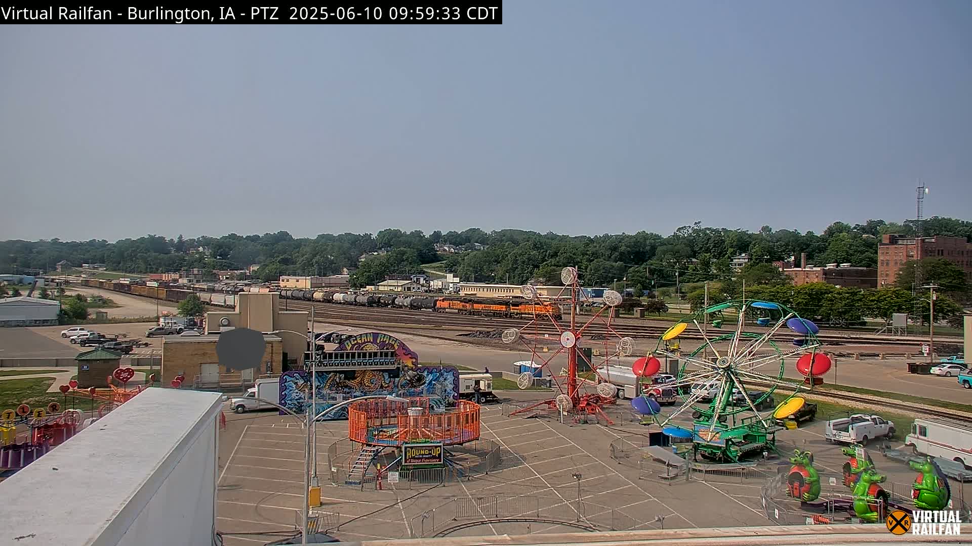 A long freight train sits on the tracks behind a carnival with several rides in a parking lot under a clear, sunny sky.